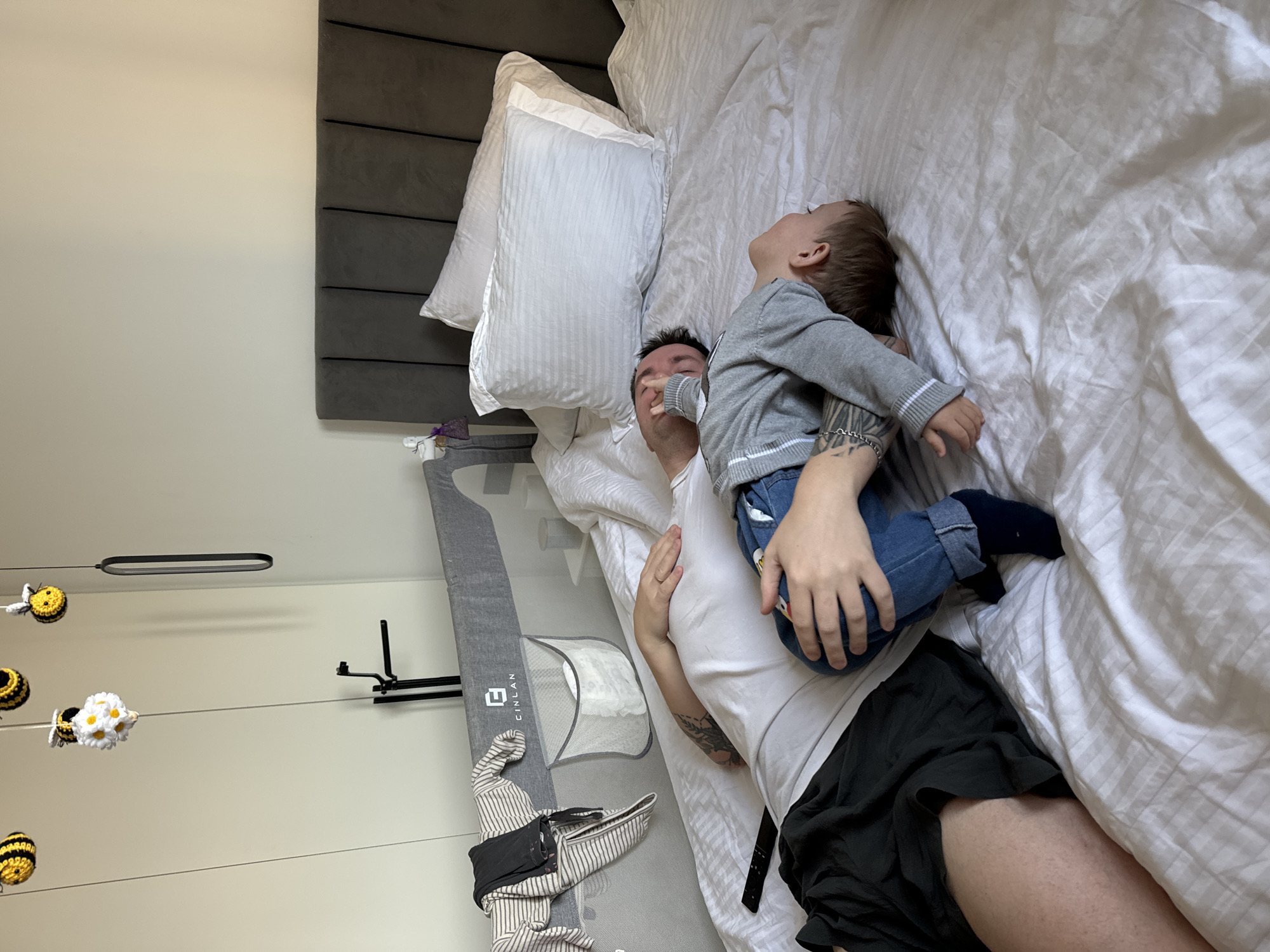 A man and a toddler are lying on a bed in a room. The man is wearing a white t-shirt and dark shorts, and the toddler is wearing a grey long-sleeved shirt and blue jeans. The toddler is looking upwards. There is a grey padded headboard behind the pillows. A portable changing pad and laundry hamper are to the left of the bed. The wall has a bee-themed mobile hanging from the ceiling. The location is Odintsovo, Russia.