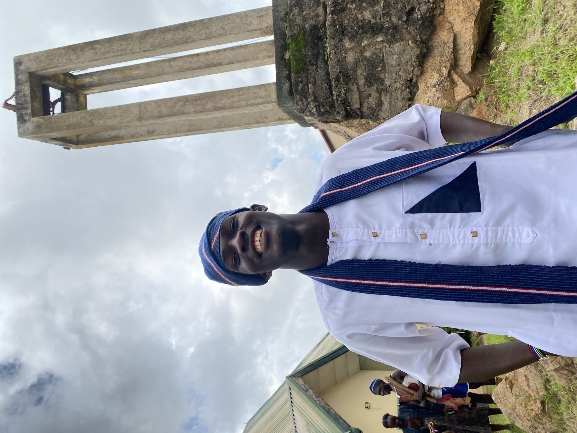 The image depicts a young Black man smiling at the camera, wearing a white tunic and a blue and white striped headwrap, in front of a stone monument. He has a blue and white striped sash draped around his neck. The monument is tall and appears to be made of concrete, with open sections near the top. In the background, to the left of the man, is a building with a green, sloped roof. To the far left, two other individuals are visible, dressed in traditional attire. The ground is covered with grass and scattered rocks. The sky is cloudy and overcast. The location is identified as Wang, Nigeria.