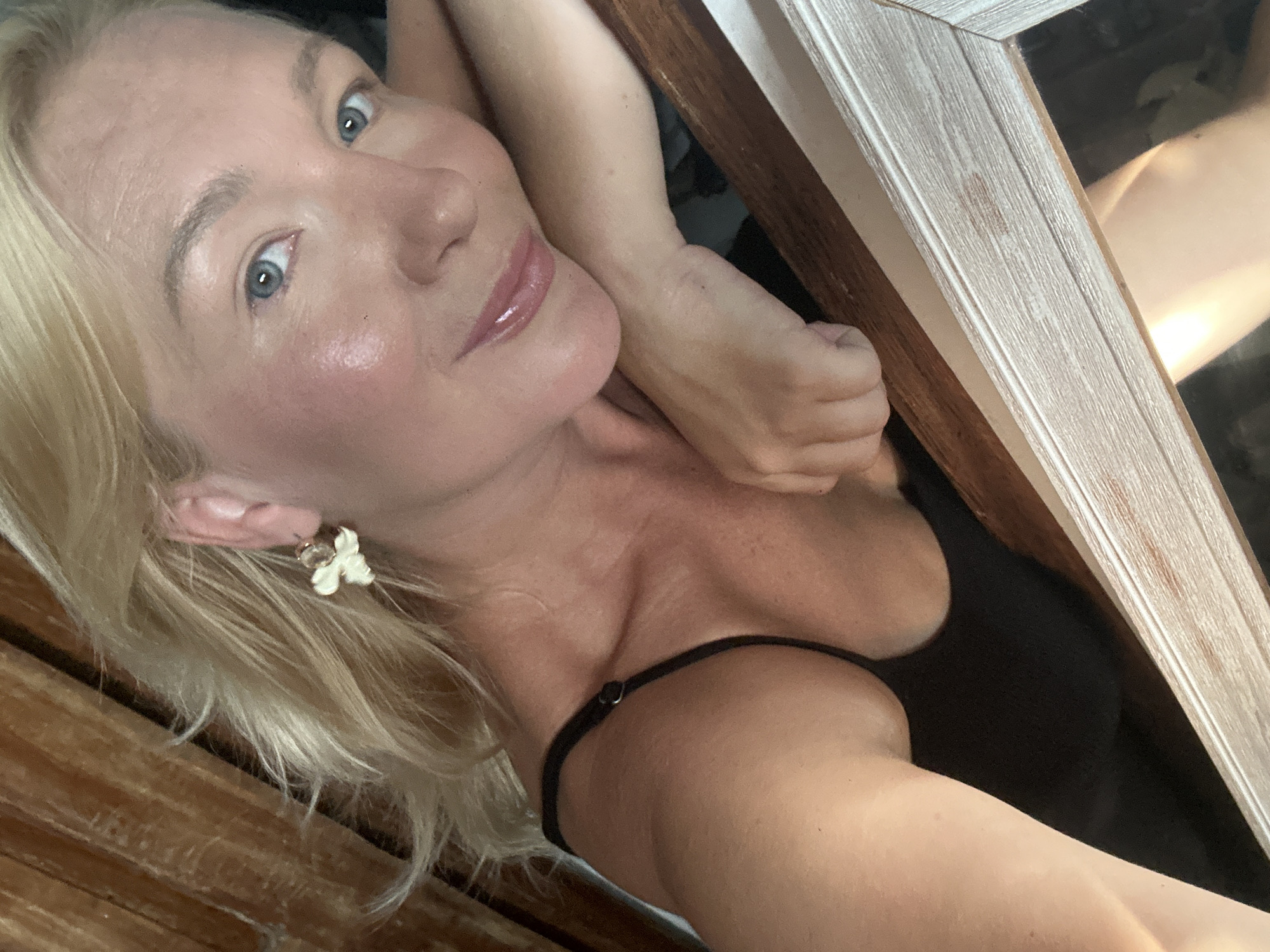 A fair-skinned woman with blonde hair poses at an oblique angle, looking into the camera. She wears a black top with thin straps and a small flower-shaped earring. She appears to be in an indoor setting. Parts of a wooden structure are visible around her. There is no evidence to suggest this is in North Kuta, Indonesia.