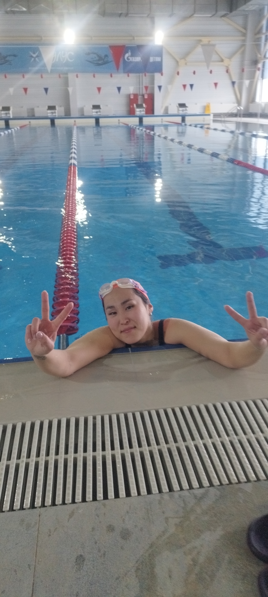 A woman with a pink headscarf and swimming goggles rests at the edge of an indoor swimming pool in Yakutsk, Russia. She is wearing a dark swimsuit and giving a peace sign with both hands. The pool is divided into lanes by red and white lane markers. The pool area includes starting blocks and a banner with logos. The floor around the pool is concrete with a white metal drainage grate.