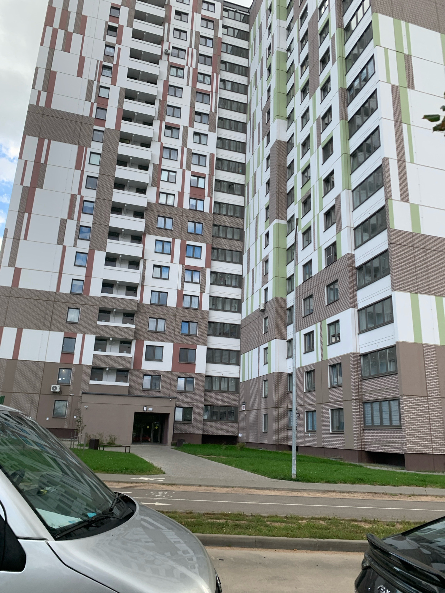 The image depicts a tall, modern apartment building in Minsk, Belarus. The building features a distinctive facade with alternating panels of white, reddish-brown, and light green stripes. Numerous windows and balconies are visible on each floor, indicating residential units. In the foreground, the hood and windshield of a silver car are prominent, suggesting it is parked on a street or in a parking area. To the right, the rear of a black car with a spoiler is partially visible. The ground in front of the building is a paved walkway leading to an entrance, with patches of green grass on either side. The sky is cloudy.