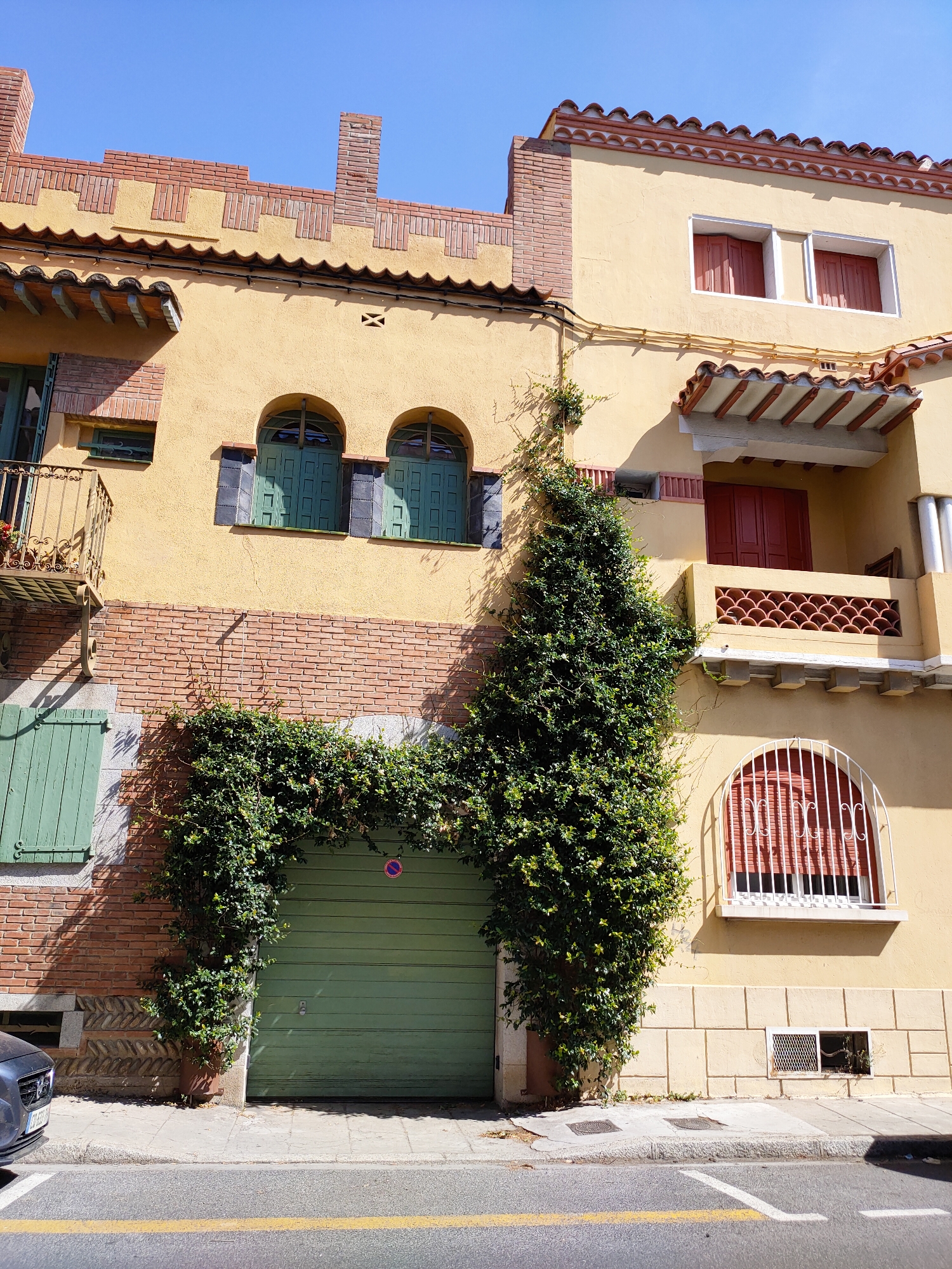 The image shows a facade of a building in Perpignan, France, bathed in sunlight under a clear blue sky. The building features a light yellow stucco exterior with decorative brickwork accents, particularly along the roofline and around windows and balconies. There are several arched windows with green shutters on the main section of the building. A balcony with an ornate metal railing juts out from the left side. A vibrant green garage door is centrally located, partially obscured by a thick growth of ivy that cascades down the wall, forming an archway over the door. The ivy also climbs the wall to the left and right of the garage. To the right of the garage, the facade continues with more yellow stucco, featuring a balcony with a patterned railing and red-shuttered windows. An arched window with a white metal grille is visible on the lower right. The street in front of the building is paved with asphalt, marked with yellow and white lines. A portion of a dark-colored car is visible on the left side of the street. The overall scene suggests a residential or commercial building in a European city.