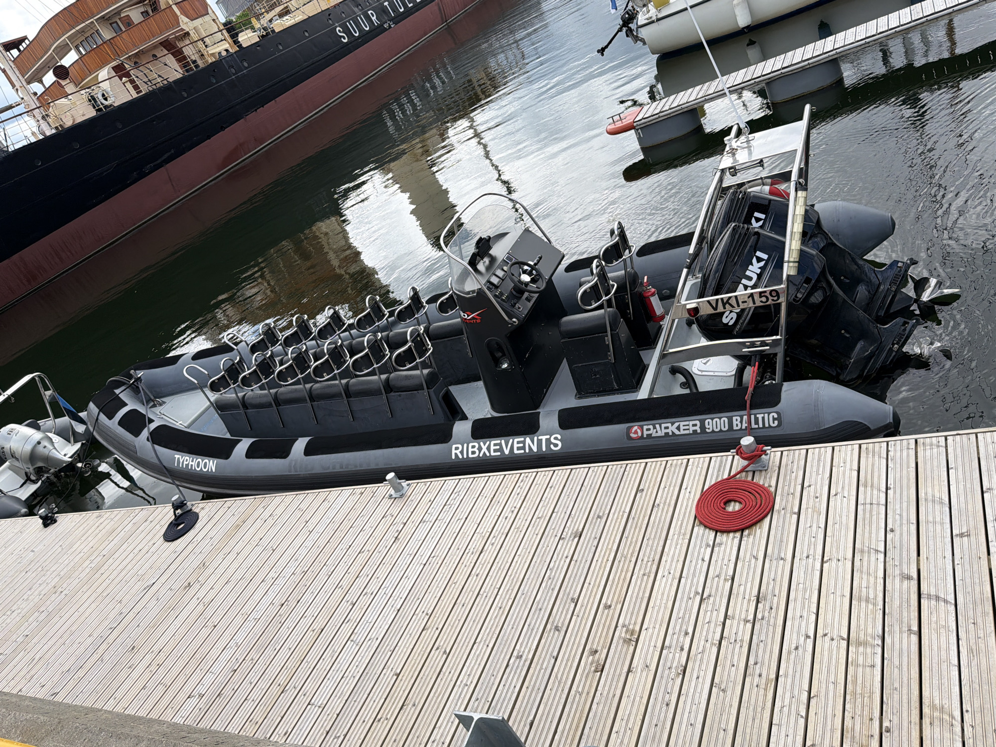 In Tallinn, Estonia, a black and grey rigid-hulled inflatable boat (RHIB) named "TYPHOON" is docked at a wooden pier. The boat has multiple rows of passenger seats with silver railings, a steering console with a windshield, and twin outboard engines at the stern. The text "PARKER 900 BALTIC" is visible on the side of the boat. A red rope is coiled on the pier, securing the boat. In the background, a large, dark-hulled ship with the partial text "SUUR TULLE" is visible, along with other docked vessels and floating pontoons. The water is calm and reflects the sky and the surrounding structures. No people are visible in the image.