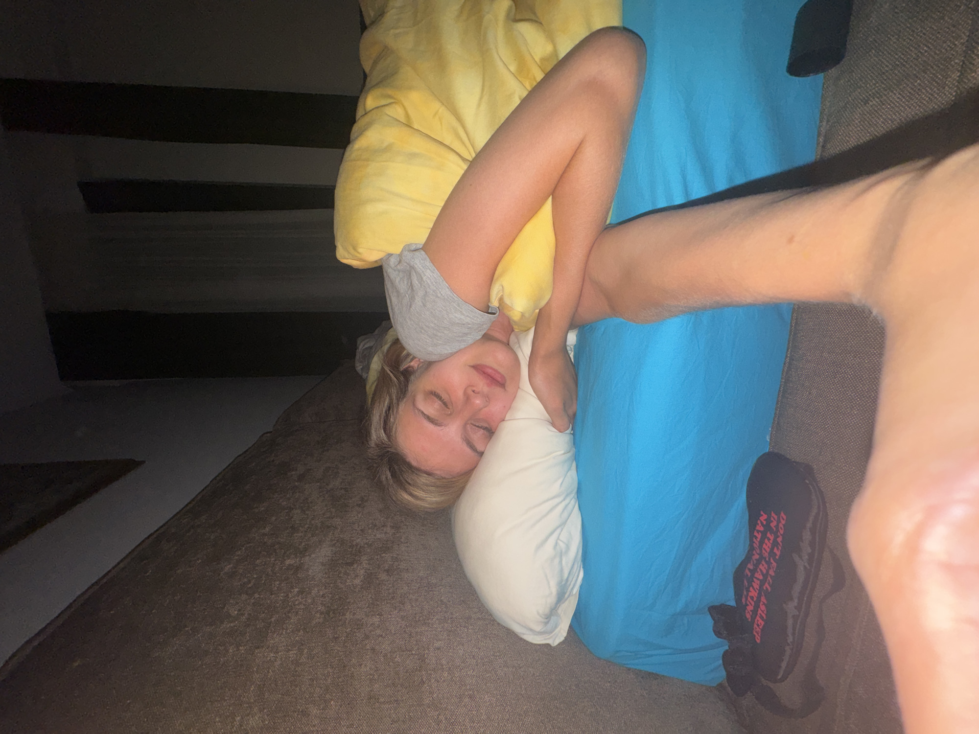 A young woman is lying on a brown sofa in a dimly lit room in an unknown location. She is under a yellow blanket and a blue sheet, with her head resting on a white pillow. The woman is wearing a gray t-shirt and appears to be asleep. A pair of black slippers are visible on the sofa next to her. The background includes a dark wall and floor.