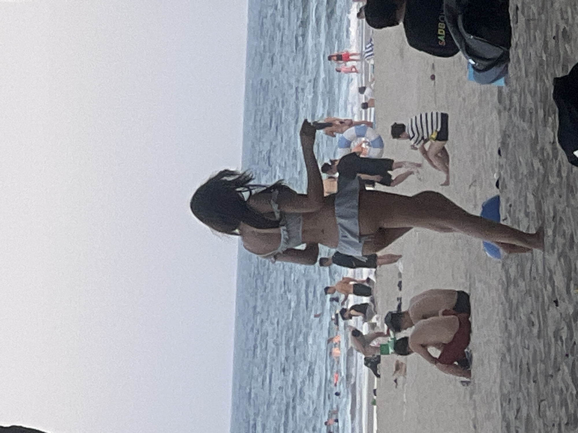 The image depicts a crowded beach scene, likely in Đà Nẵng, Vietnam, given the commonality of such scenes there. The sky is overcast, and the sea is a deep blue with moderate waves. In the foreground, a woman in a light blue bikini with ruffled trim is walking away from the camera towards the water. She has long, dark hair.
On the beach, there are numerous people scattered around. To the left of the woman in the foreground, two men are sitting on the sand. One is wearing red shorts and the other black shorts. Further back, a group of people are visible near the water's edge, some wading, others standing. There are also several beach umbrellas and towels.
In the middle ground, more people are present. A man in an orange shirt stands with another man in a black shirt and shorts. A person in a striped shirt and black shorts is sitting further back. In the far distance, the water stretches to the horizon. The overall atmosphere is one of a busy summer day at the beach.