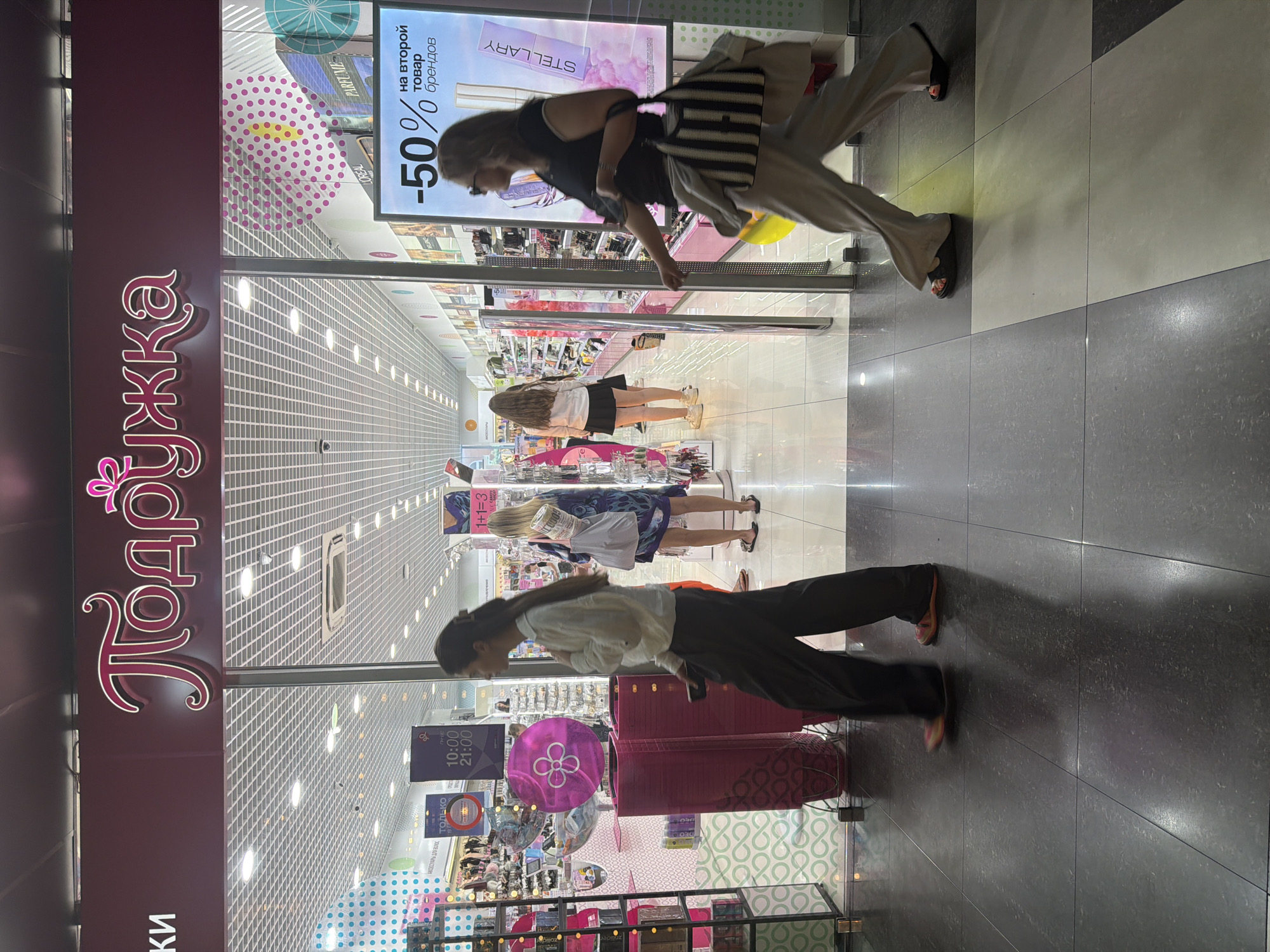 The image depicts the exterior of a "Podruzhka" cosmetics store, indicated by the prominent Russian text on the signage. The store is brightly lit, suggesting it is open for business. Inside the store, shelves are stocked with various beauty products, hinting at a retail environment focused on cosmetics and personal care.
Several individuals are visible in the image. One woman, wearing glasses and a black top, is walking past the store. Another woman, dressed in a white blouse and dark pants, is looking into the store. A third woman, in a blue patterned dress, is also browsing the merchandise inside. The floor tiles suggest an indoor mall or shopping center setting.
A prominent advertisement displays "-50% on the second item from brands," indicating a sale is currently in progress. Other visible text includes store hours: "10:00 - 21:00" and "Only 1+1=3." The overall atmosphere appears to be a typical day at a shopping venue, with people engaged in browsing and shopping for beauty products. The lighting and indoor setting imply the time of day is likely within business hours, and the weather is not a factor as it is an interior shot. The location is identified as Sochi, Russia.