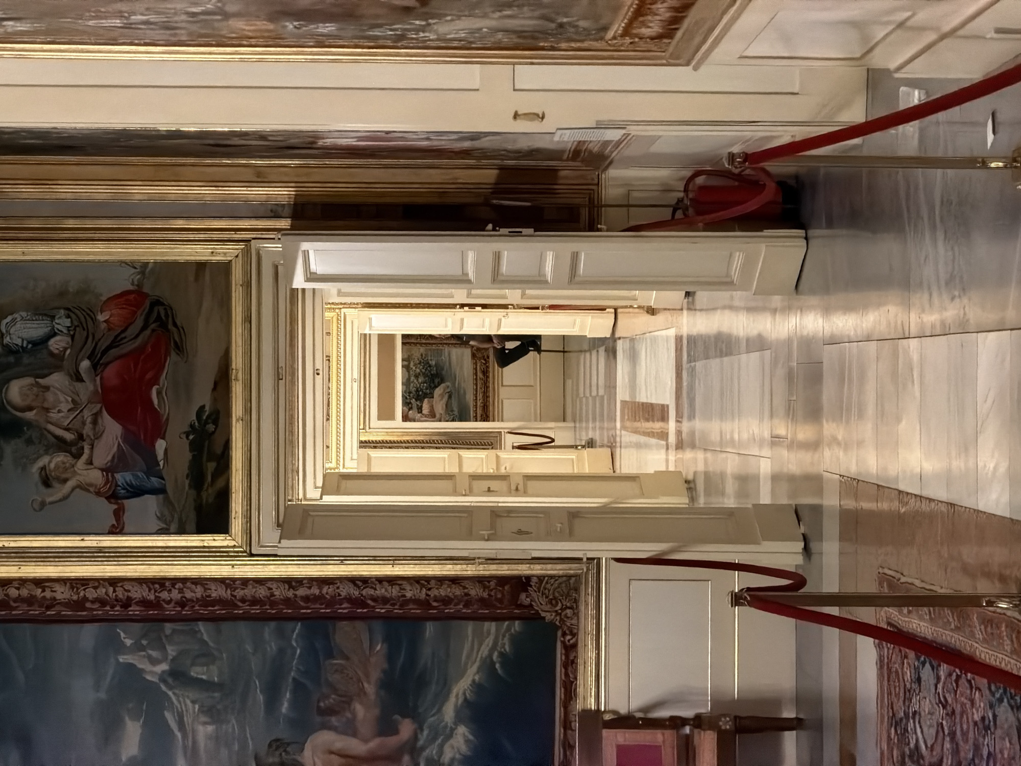 The image depicts a long, ornate hallway within a grand building, likely a palace or museum. The hallway is lined with large, elaborately framed paintings displayed on the walls. The architecture features classic design elements, including tall, white-paneled doors and high ceilings with decorative moldings. Sunlight streams in from unseen windows, casting long shadows and highlighting the polished marble floors, creating a dramatic and somewhat disorienting perspective due to the camera's vertical orientation.
In the foreground, a large, dark-framed painting is partially visible, featuring what appears to be a mythological or biblical scene with dramatic lighting and figures. To the left, another framed artwork shows figures in rich red and blue robes, possibly a religious depiction. Further down the hallway, more paintings are visible, receding into the distance, creating a sense of depth and grandeur.
A red velvet rope barrier, supported by brass stanchions, runs along the right side of the hallway, guiding visitors and indicating that the area may be an exhibition space. In the mid-ground, a fire extinguisher is mounted on the wall near a doorway, a practical safety detail within the opulent setting. The overall impression is one of historical significance and artistic wealth, likely within a well-preserved cultural heritage site. The lighting suggests it is daytime, and there are no indications of weather. The scene evokes a sense of quiet contemplation and admiration for the art and architecture.