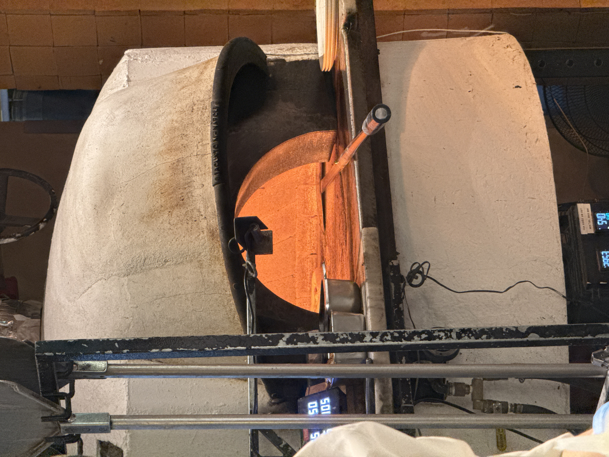 The image shows a traditional wood-fired pizza oven in a restaurant. The oven is made of brick and has a dome-shaped roof. The opening of the oven is visible, with a pizza peel inside, suggesting that a pizza is being cooked or is about to be removed. The interior of the oven glows with orange flames. To the left of the oven, there are metal racks and a digital display showing numbers. In the background, stacks of plates are visible on a counter. The overall scene is characteristic of a pizzeria, likely in Hà Nội, Vietnam, given the typical presence of such ovens in restaurants serving Italian cuisine.