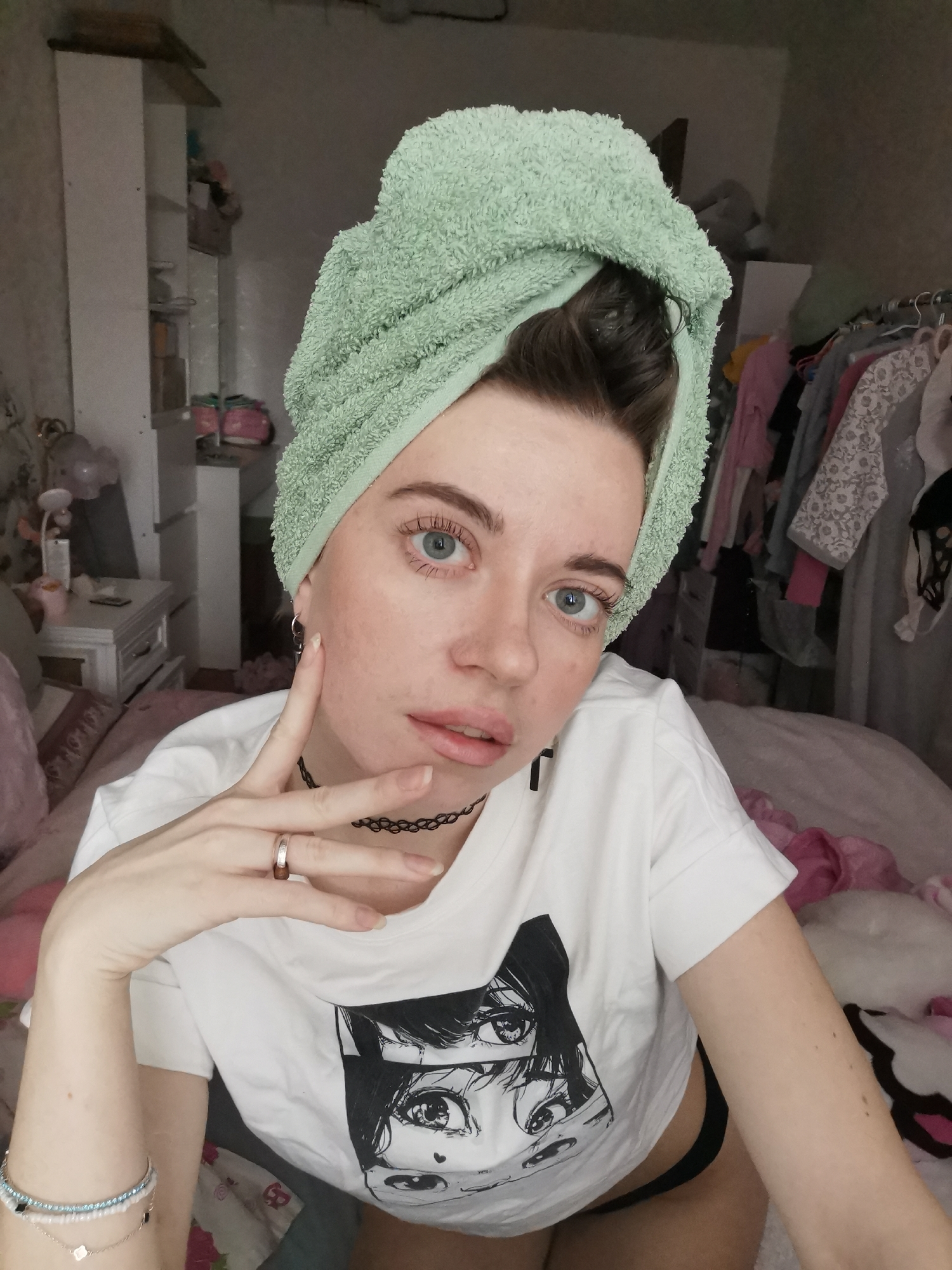 A young woman is captured in a close-up selfie indoors, likely in a bedroom. She wears a light green towel wrapped around her head, indicating recently washed hair. Her blue eyes look directly at the camera, and her expression is a neutral, slightly pouting pose. She has fair skin, with visible freckles or minor blemishes.
She is dressed in a white short-sleeved t-shirt featuring a prominent black and white graphic print of large, expressive eyes in an anime or manga style. Around her neck is a black choker necklace. On her right hand, she wears a simple ring on her middle finger, while her left wrist is adorned with multiple delicate bracelets. Her right hand is raised near her face, with two fingers slightly parted in a gesture.
The background reveals a personal and somewhat cluttered room. To the left, a white shelving unit or tall cabinet is visible, stocked with various items. Adjacent to it, a white nightstand holds a lamp and other small objects. To the right, a rack of clothes on hangers fills part of the frame, and a pink and white bed or bedding is partially seen. A white, possibly Hello Kitty-themed, pillow or object is also visible on the bed. The lighting is soft and diffused, characteristic of an indoor setting without direct, bright natural light. No explicit text is visible in the image, apart from the graphic on her t-shirt.