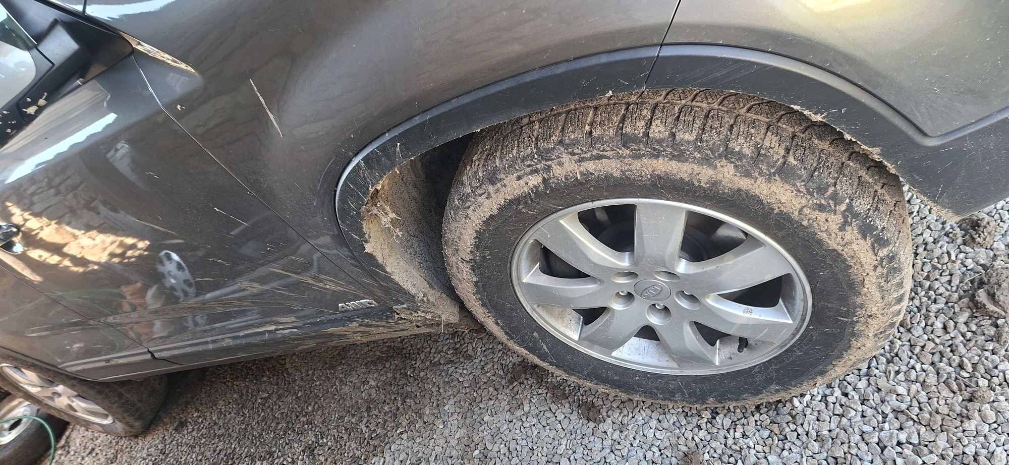 The image displays the side profile of a dirty, dark gray Kia SUV, parked outdoors on a gravel surface. The vehicle is heavily soiled with mud and dust, particularly on the front passenger-side tire, its alloy wheel, and the surrounding wheel well and lower body panels. The tire exhibits a distinct, chunky tread pattern, typical of all-season or winter tires, coated in dried mud. The silver alloy wheel prominently features the "KIA" logo on its hubcap.
Evidence of recent travel through muddy terrain is clear from the significant dirt splatters and streaks across the car's paintwork, extending from the tire up the fender and door panel. Several visible scratches, including a distinct white line on the fender and smaller marks near the door handle area, suggest wear and tear. Reflections on the car's glossy surface reveal glimpses of a nearby structure, possibly a building with brickwork, and bright daylight conditions.
The ground beneath the car is composed of small, light gray gravel stones, interspersed with patches of accumulated mud and dirt. A partial view of another tire or wheel is visible in the bottom left corner, suggesting another vehicle or a spare wheel. The "AWD" badge is clearly visible on the lower part of the car's side panel, indicating its all-wheel-drive capability. The scene captures the vehicle in a state reflecting rugged use or recent off-road driving.