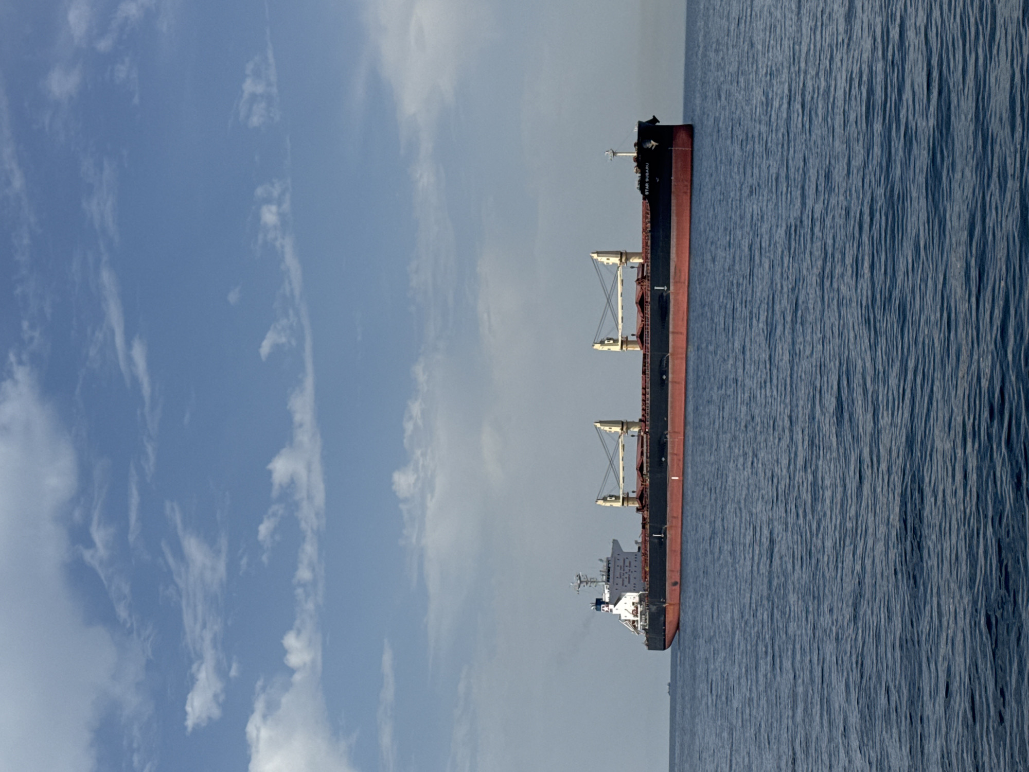 A large cargo ship, identified as the "STAR SUBARU," is sailing on a body of calm, blue water under a partly cloudy sky. The ship has a dark hull with a red waterline. It features a white superstructure towards the front, with several decks and windows. Two large yellow cranes are visible on the deck of the ship. The scene suggests an open body of water, likely the sea, with a distant horizon. The presence of a cargo ship indicates it is involved in maritime transport, possibly near a port or a shipping lane. Considering the provided location of Marina di Ravenna, Italy, this image likely depicts the ship at sea off the coast of this Italian port city.