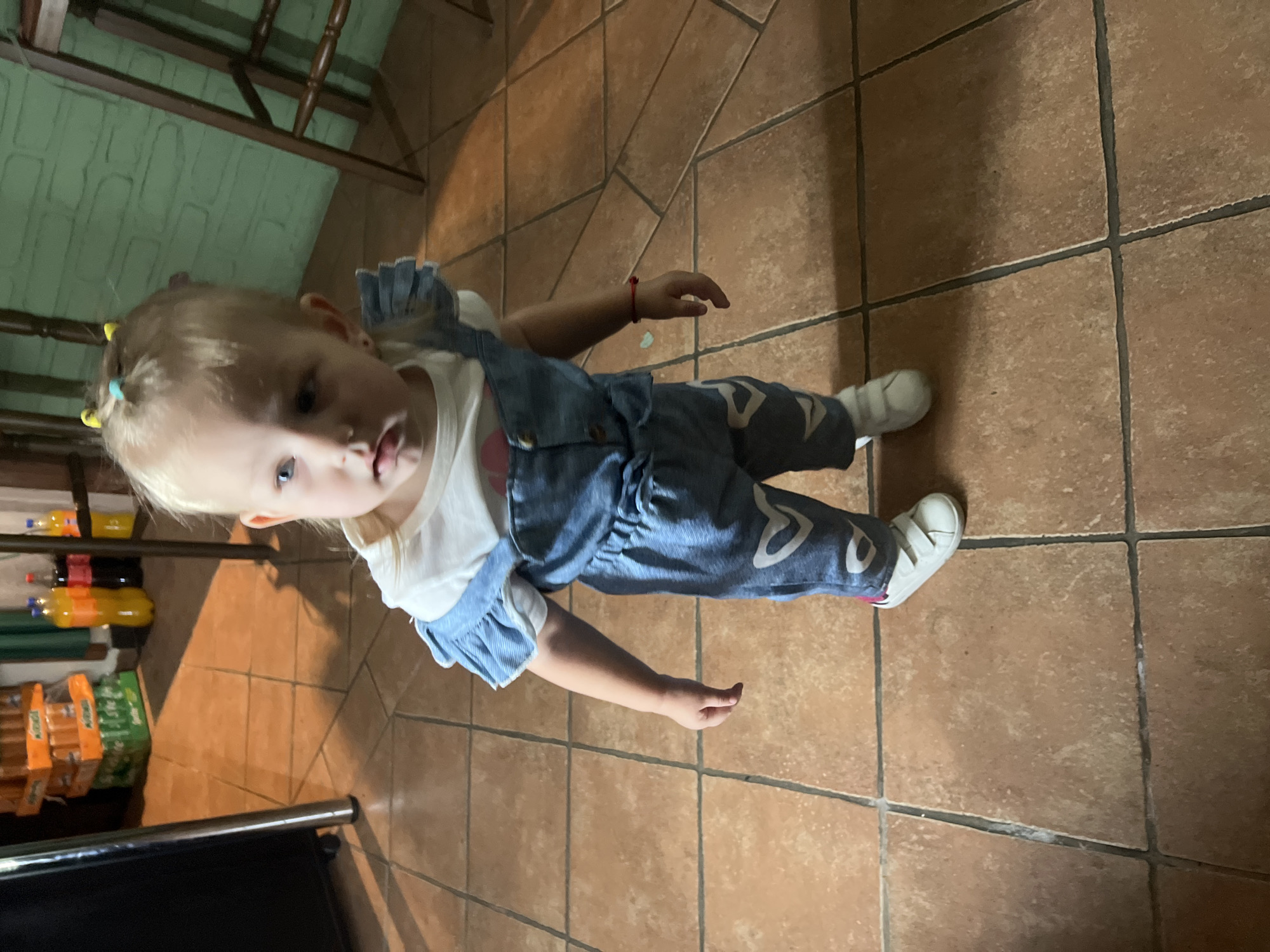 A blonde toddler girl with blue eyes is standing on a tiled floor. She is wearing a white t-shirt and denim overalls with hearts on them. She has yellow hair ties in her hair. In the background, there are bottles of drinks stacked on shelves and a wooden chair. The environment appears to be indoors, possibly a kitchen or dining area, with a green brick wall visible on the right side of the image. The lighting suggests it is daytime.