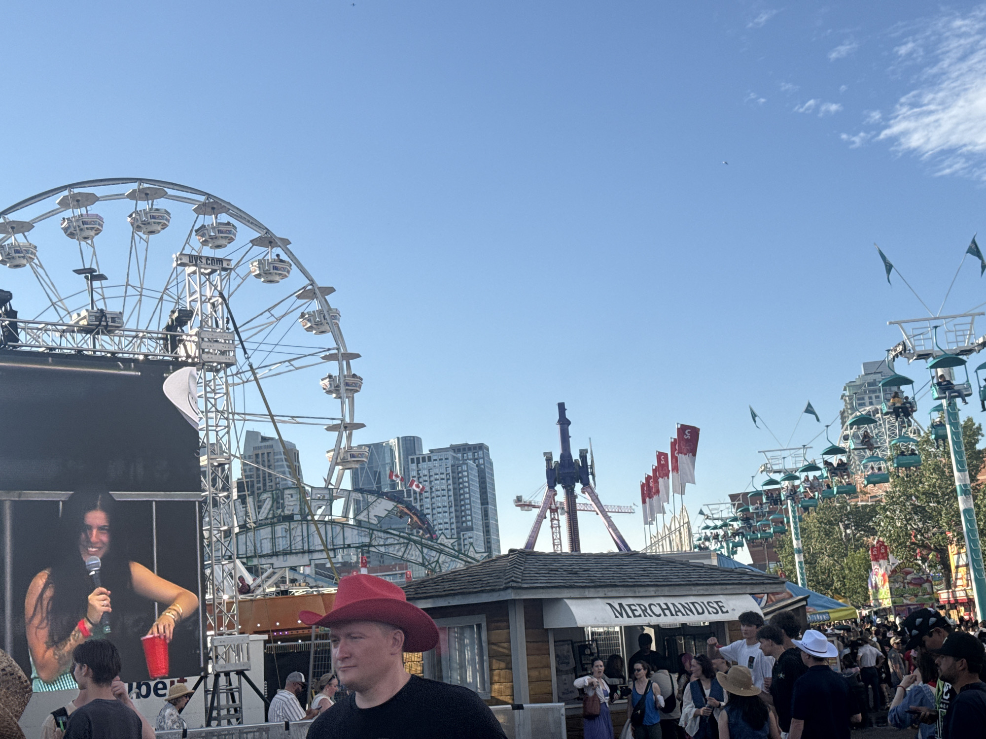 The image depicts a busy outdoor event, likely a fair or festival, with various amusement rides and attractions visible. In the foreground, a man wearing a red cowboy hat is looking to the left of the frame. Behind him, a "MERCHANDISE" booth is visible, with several people browsing or interacting with the vendors. Further back, a large Ferris wheel and other rides dominate the background, set against a backdrop of tall buildings, suggesting an urban environment. The presence of Canadian flags on some structures, along with the distinctive architecture of the Calgary Tower and other downtown buildings, confirms the location as Calgary, Canada. The scene is bathed in the warm light of a sunny day, with a clear blue sky overhead. A large screen on the left side of the image shows a woman holding a microphone and a red cup.