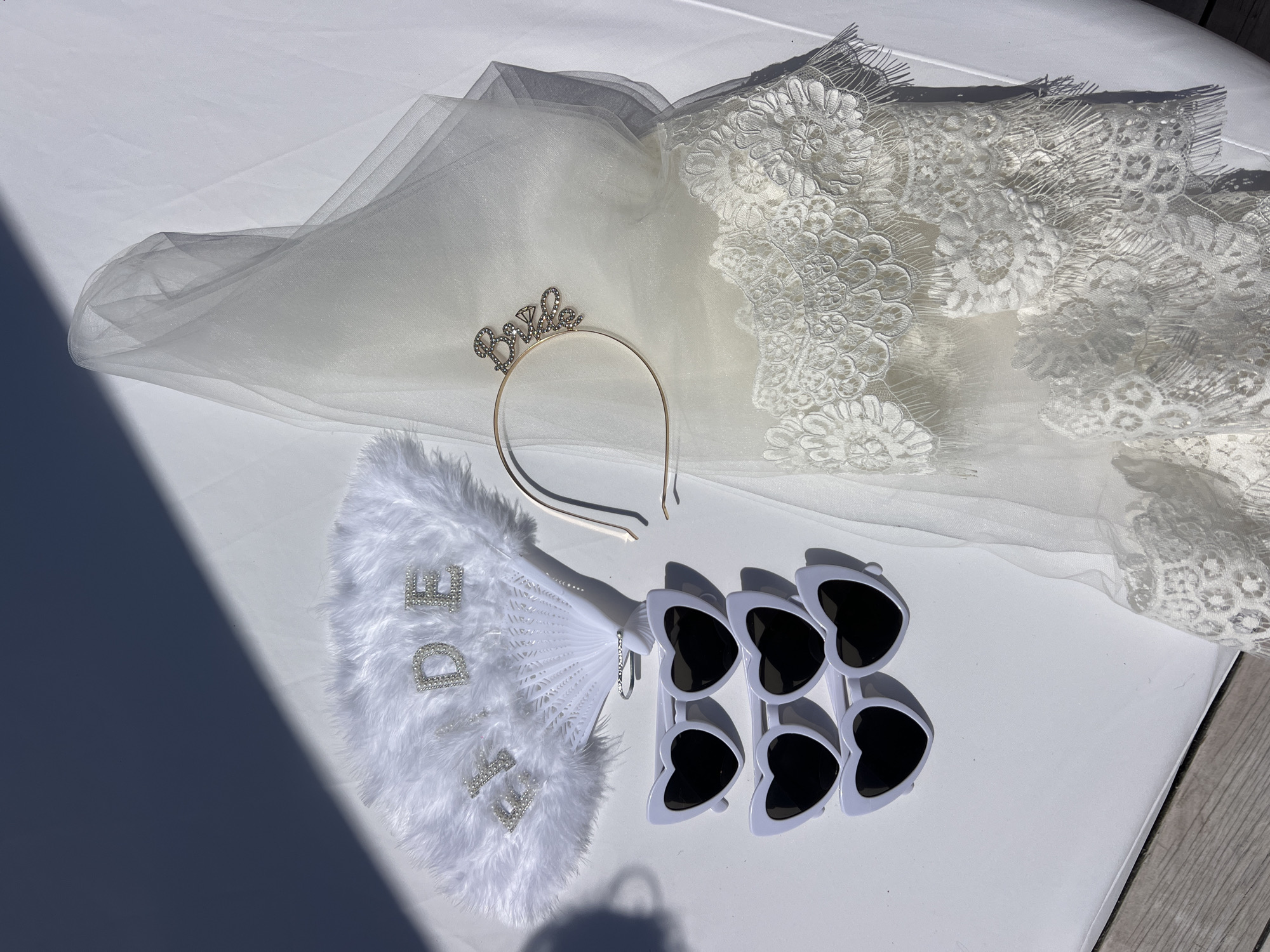 In Odesa, Ukraine, on a sunlit white surface, a collection of bridal party accessories is arranged. To the left, a white feathered fan with "BRIDE" spelled out in rhinestones is positioned. Below the fan are four pairs of white heart-shaped sunglasses with black lenses. To the right, a sheer white veil with lace trim is draped, and a gold headband with the word "Bride" and a diamond accent is placed on top of the veil. The scene is illuminated by natural light, casting shadows from an unseen object across the surface.