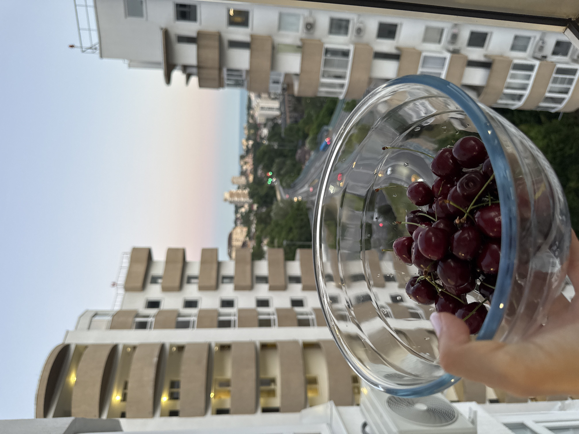 A person's hand holds a clear glass bowl filled with ripe cherries. The bowl is positioned in the foreground, with a soft focus on the fruit and the hand. In the background, a cityscape unfolds under a twilight sky. Two tall residential buildings, characterized by cream-colored facades and curved balconies, dominate the mid-ground. Between these buildings, a winding road with car headlights and taillights is visible, suggesting it's dusk or early evening. Further in the distance, more buildings and a hint of the Black Sea can be seen. The overall scene suggests a view from a high-rise apartment in Sochi, Russia, likely a residential area.