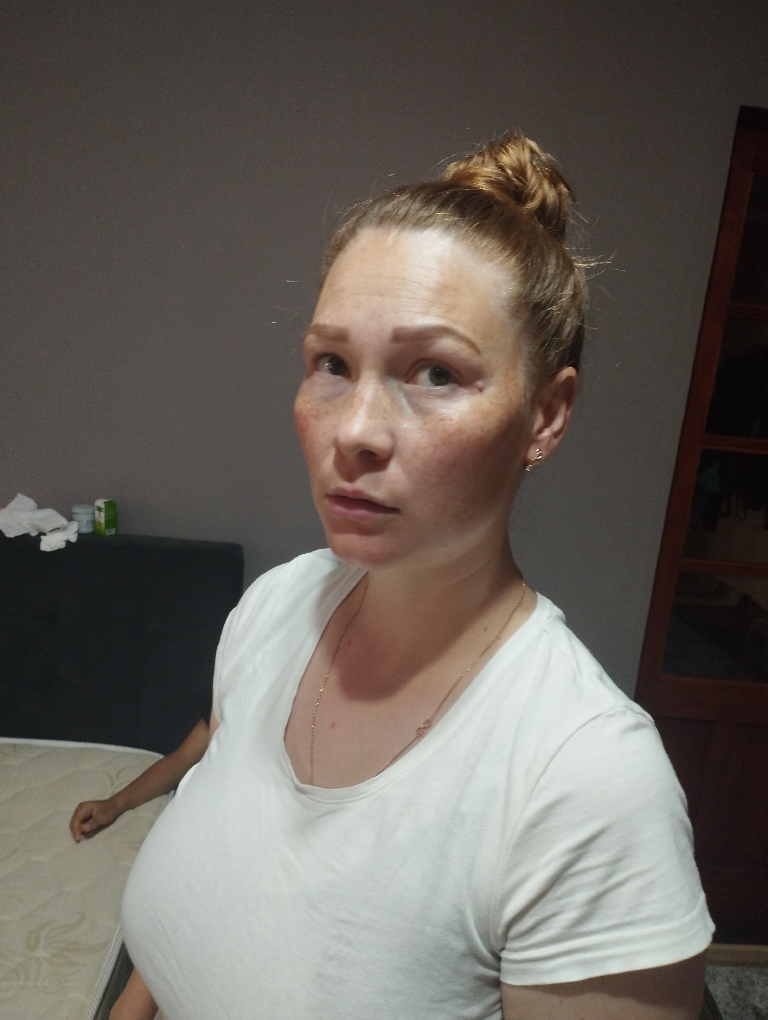 The image shows a woman in a white t-shirt with her hair in a bun. She has freckles on her face. She is looking to the side. In the background, there is a wall, a mattress, and what appears to be a cabinet or door. The lighting suggests an indoor setting. There are no visible indications of the location being Fălești, Moldova.