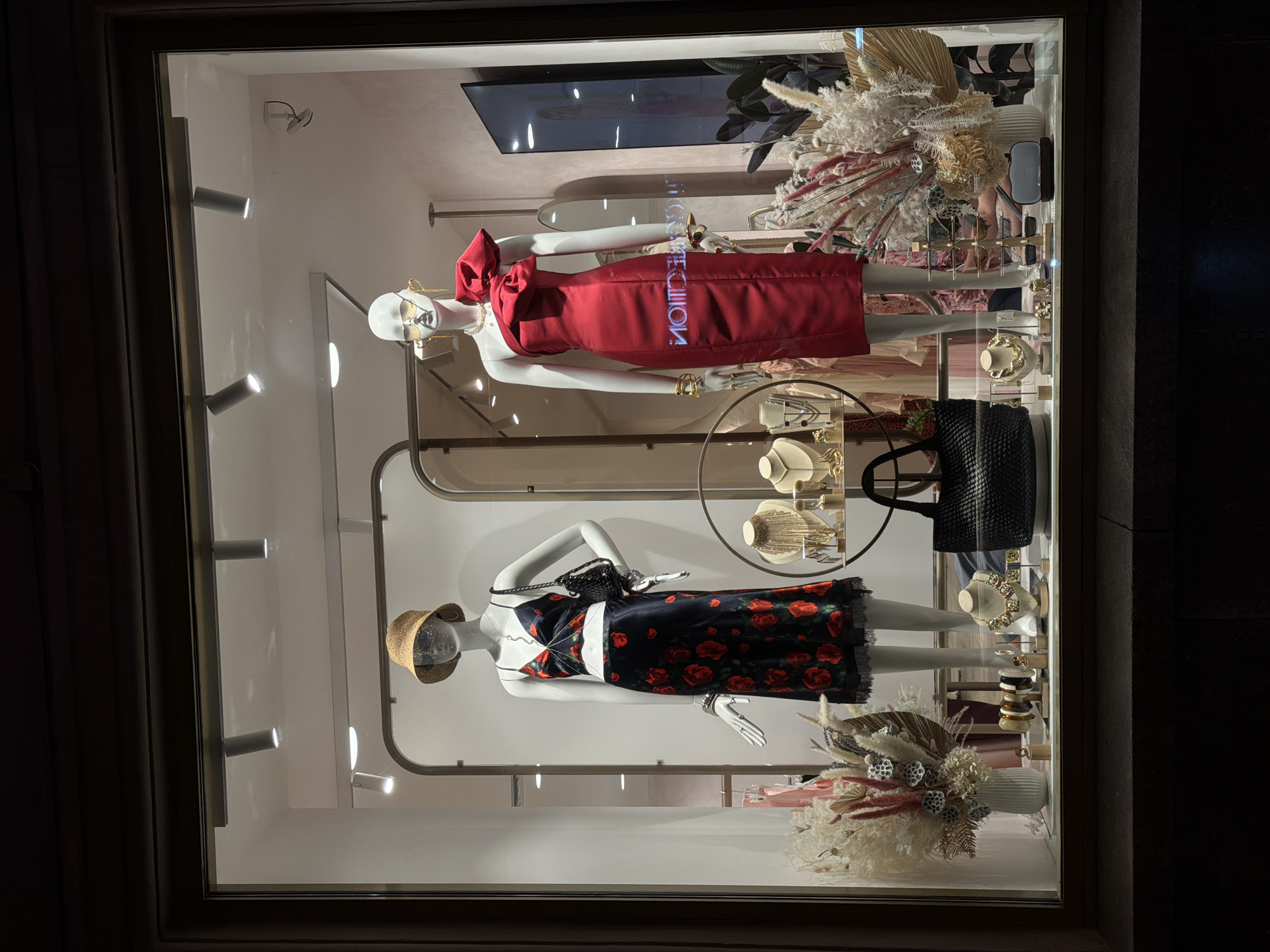 A storefront window display in Moscow, Russia. Two mannequins are the focal point. The top mannequin, facing left, wears a red dress with a large bow and sunglasses. The bottom mannequin, also facing left, wears a black and red floral dress and a straw hat. In between the mannequins is a display of jewelry and a black handbag. Decorative dried floral arrangements are present at the top and bottom of the window. Track lighting fixtures illuminate the scene.