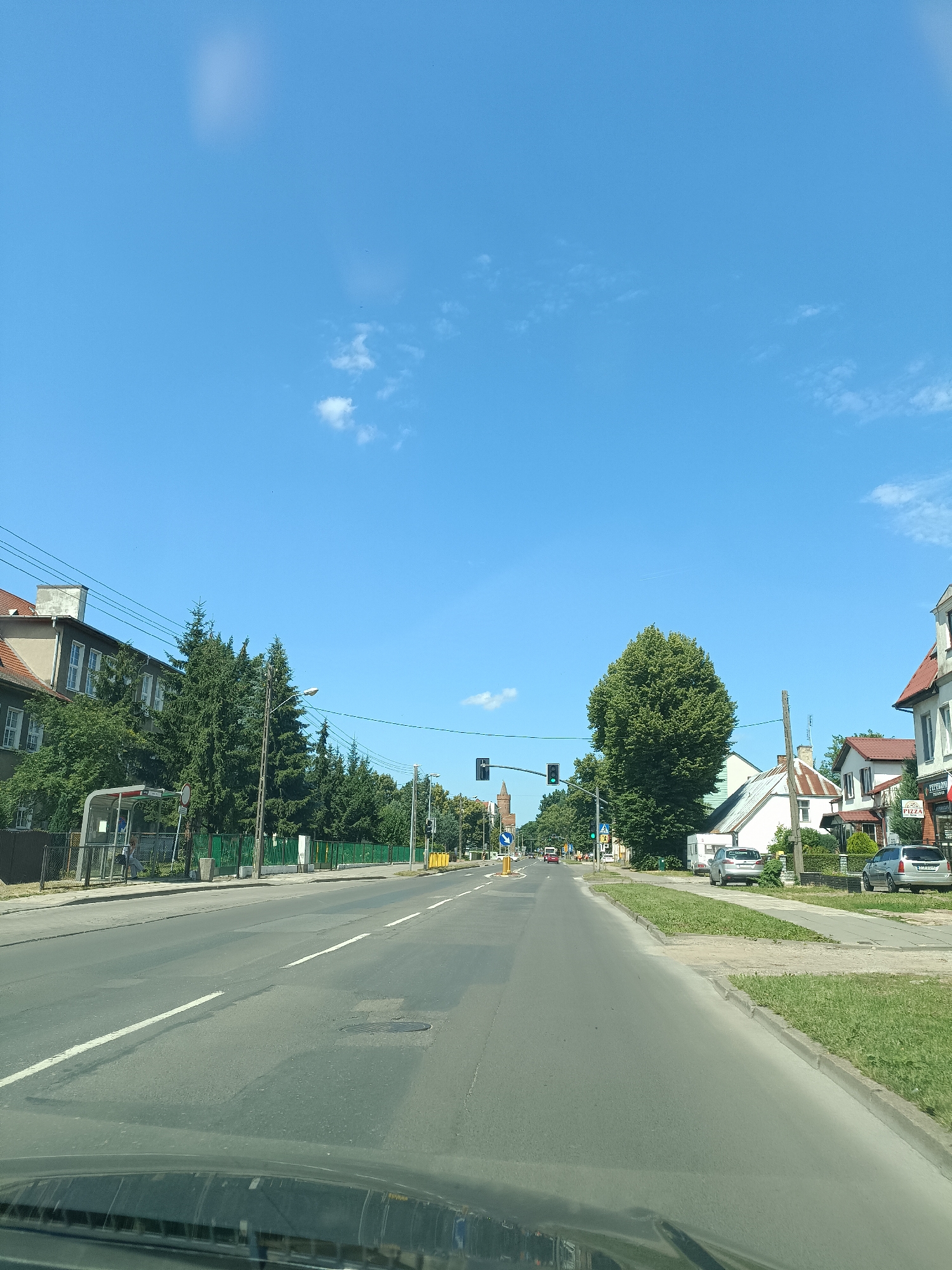 The image depicts a street scene in Gryfino, Poland, under a clear blue sky with a few wispy clouds, suggesting a sunny daytime. The perspective is from a vehicle driving down a paved road with painted lane markings.
On the left side of the road, there is a bus stop with a shelter and a person sitting inside. Adjacent to the bus stop is a building with a red-tiled roof and several windows, surrounded by tall evergreen trees. Further down the road, traffic lights are visible, with the one on the left showing green.
On the right side of the road, there are several houses and buildings. One building has a sign that reads "PEPPERONI PIZZA". The houses have pitched roofs, and some have well-maintained gardens. There are several cars parked along the side of the road. In the distance, a tall, slender tower or spire is visible. The overall impression is a peaceful suburban or small-town street on a pleasant day.