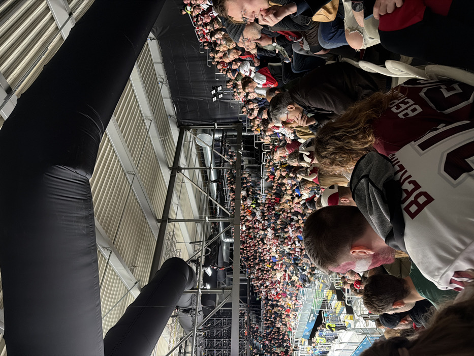 A crowded stadium view from a high vantage point. People are seated in rows. A large, black inflatable structure is on the left. Metal support beams and stadium infrastructure are visible. Some individuals wear sports jerseys. The orientation of the image is sideways.