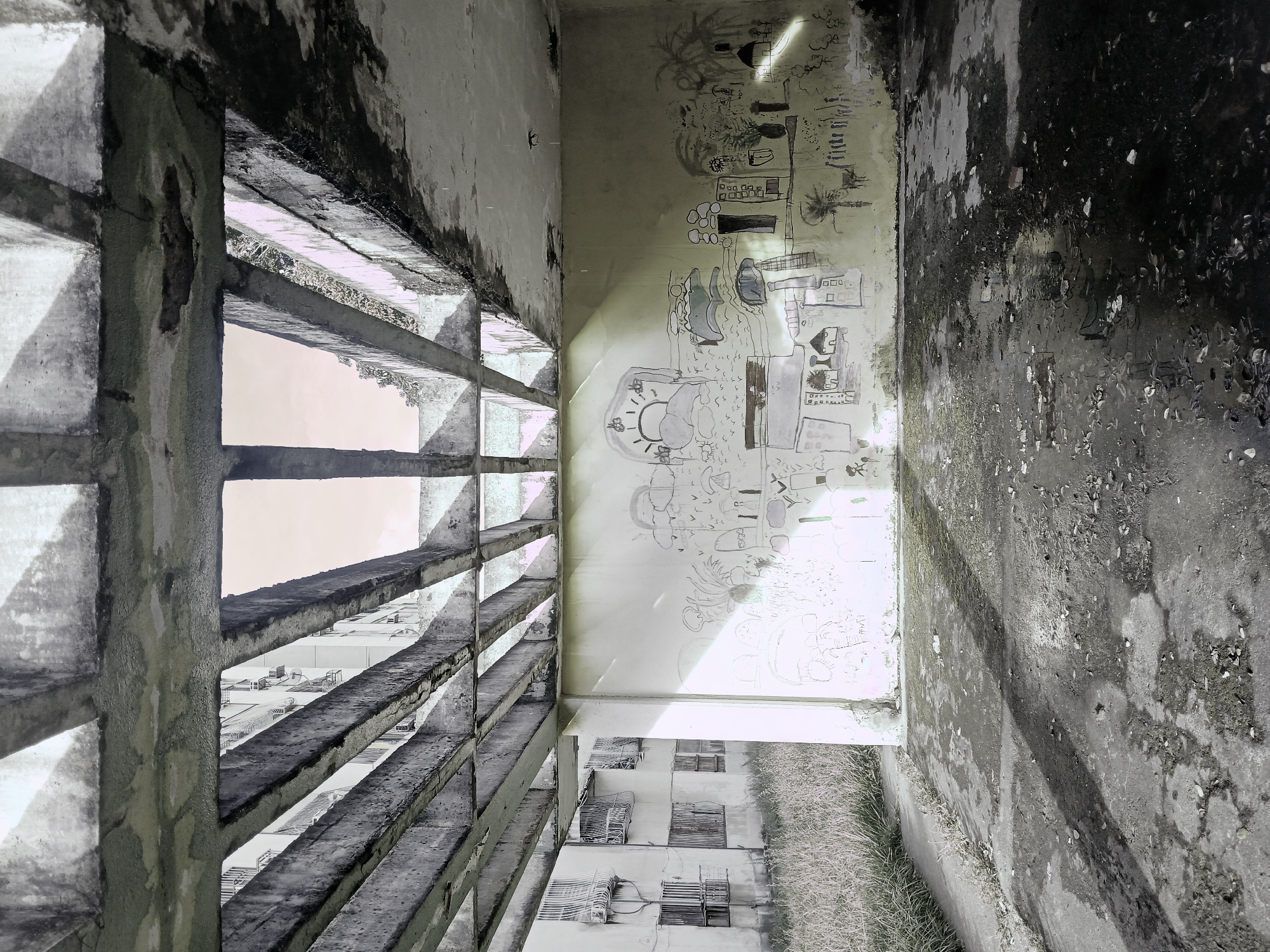 The image depicts an abandoned, open-air structure with a concrete ceiling supported by parallel concrete beams. Sunlight streams through the gaps in the beams, casting angular shadows on the ground and the wall. The floor is damp and littered with debris. The wall is covered in crude, black-and-white drawings that appear to be children's art, featuring a sun, clouds, houses, trees, and stick figures. To the left, a grassy area borders a concrete path, and beyond that, a multi-story apartment building with barred windows stands. The overall scene suggests neglect and decay in an urban environment.