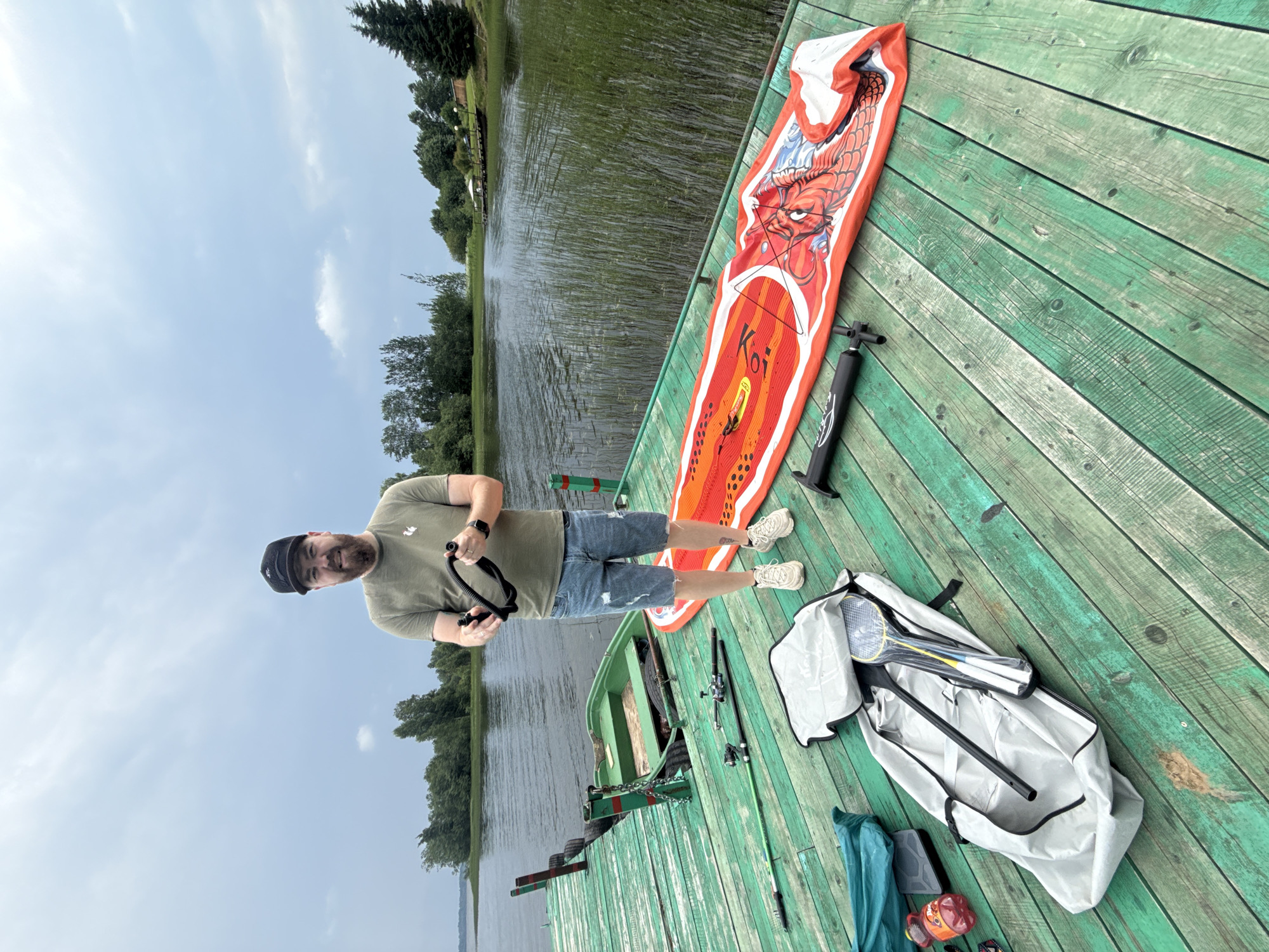 A man stands on a weathered green wooden dock by a lake in Nikol'skoe, Russia. He wears a green t-shirt, denim shorts, a black baseball cap, and white sneakers. He holds a black flexible hose in his hands. In front of him on the dock are several items: a deflated red and orange stand-up paddleboard with a dragon design, a black air pump, a large grey bag containing badminton rackets and a paddle, a green duffel bag, and two fishing rods. A small green rowboat is partially submerged near the dock. The lake is calm with reeds growing along the bank, and trees line the distant shore under a cloudy sky.