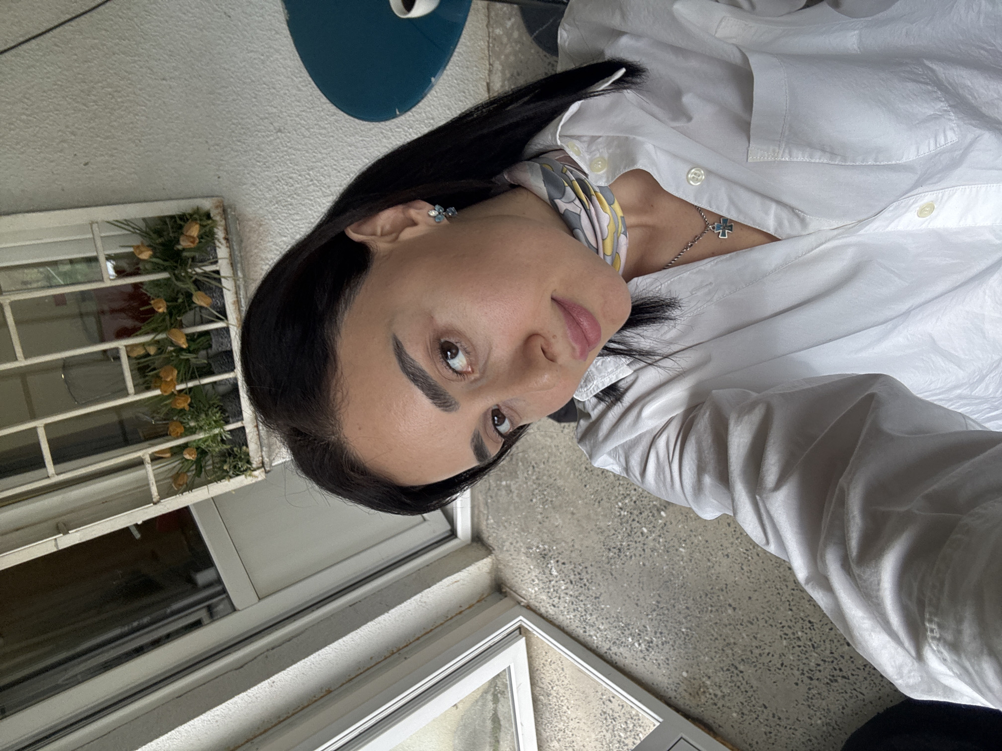 A woman poses for a selfie outdoors. She has dark hair and eyebrows, and is wearing a white button-down shirt, a scarf, and a necklace with a cross pendant. There's a window with a metal grate behind her, and some flowers. The location appears to be a balcony in Tbilisi, Georgia. The background suggests a cityscape.