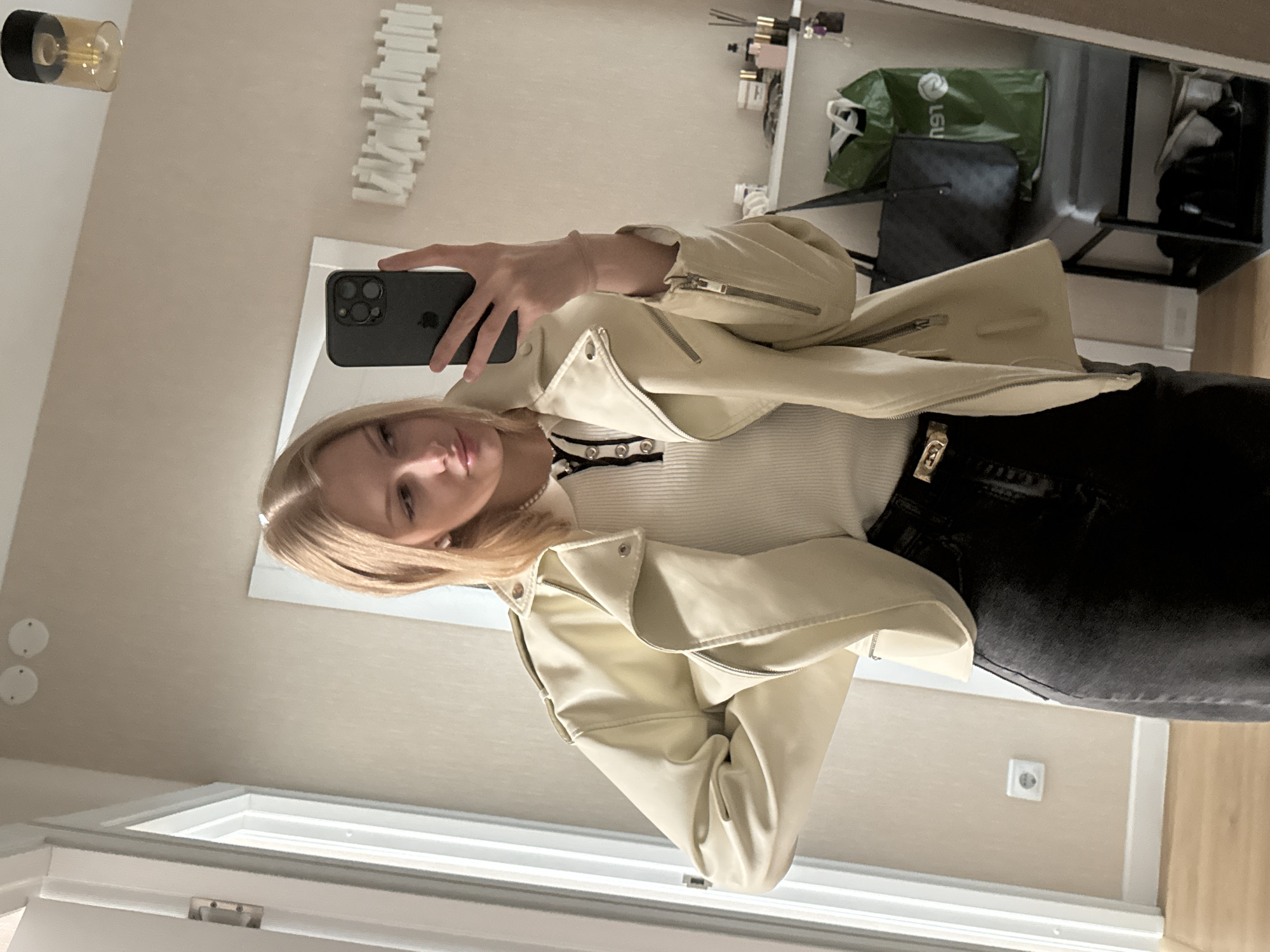 A young Caucasian female with blonde hair is depicted taking a mirror selfie in an interior room in Perm, Russia. She is wearing a light cream-colored faux leather biker jacket, a white ribbed polo shirt with a black collar and silver buttons, black jeans, and a gold-colored belt buckle. She accessorizes with pearl earrings and a pearl necklace. Her left hand is placed on her hip, while her right hand holds a dark-colored iPhone with an Apple logo visible, angled towards the mirror to capture the image.
The background, reflected in the mirror, reveals a light beige wall with a white, abstract, horizontal wall art piece on the right side. Above, two white circular ceiling fixtures are visible on the wall, and a modern ceiling light with a black base and amber-tinted glass shade hangs further right. A white floating shelf on the right holds various cosmetic items, including bottles and diffusers. Below the shelf, a grey upholstered bench or shoe rack is visible, on which a dark tote bag and a green shopping bag (with inverted text appearing to be "ЛЕНТА" / Lenta) are placed. To the left, a white door frame and part of a white door with a metallic hinge are visible. A white electrical outlet is situated on the lower left wall, above a light wooden floor.