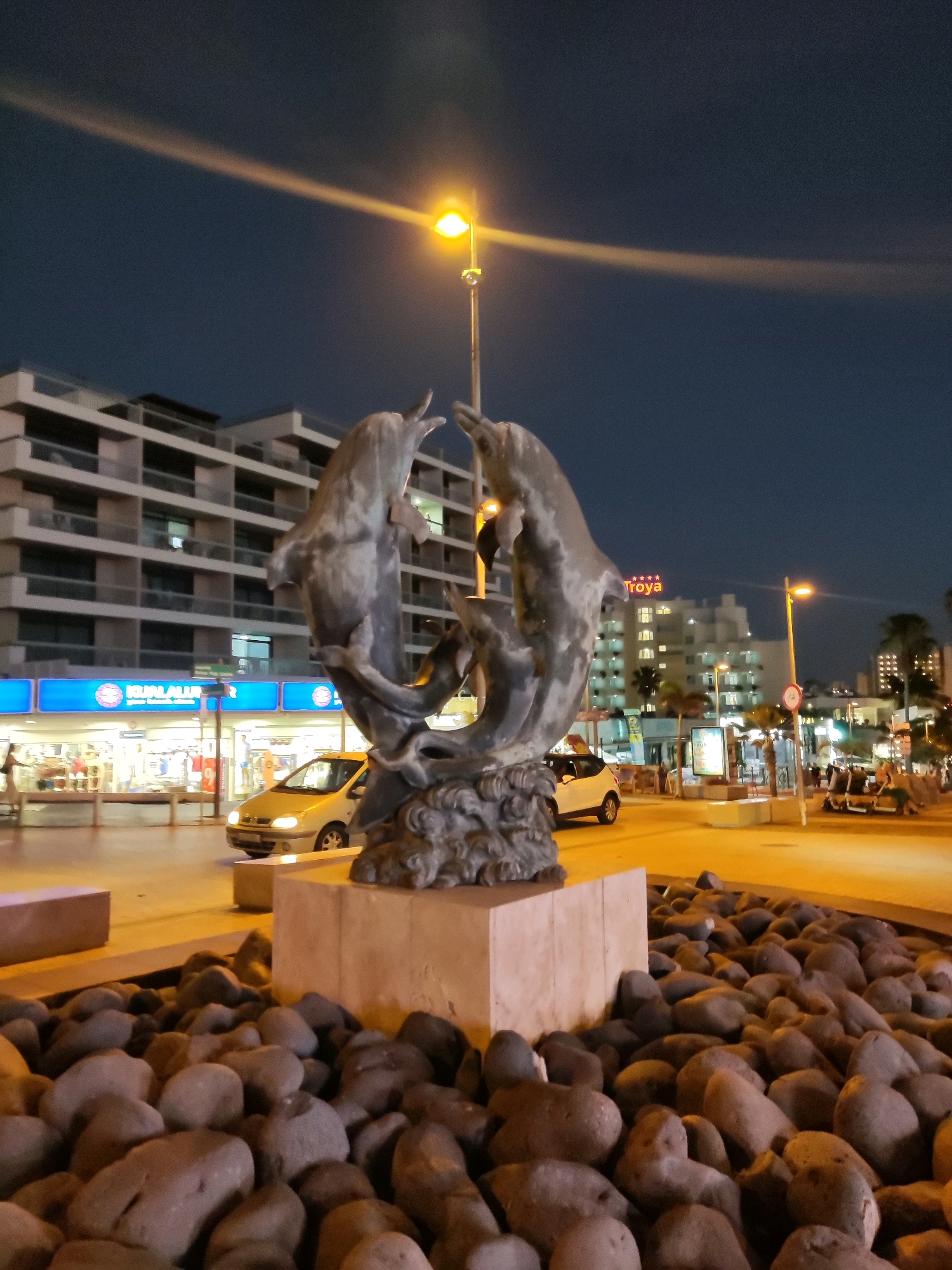 The image captures a night-time view of a dolphin sculpture in Adeje, Spain, likely situated in a public area such as a promenade or plaza. The sculpture, made of a dark material, features two dolphins in a dynamic pose, appearing to leap or intertwine. It is mounted on a stone pedestal surrounded by a bed of large, dark, rounded stones. In the background, a multi-story building with balconies and illuminated windows suggests a hotel or residential complex. Across the street, more buildings are visible, including one with a sign that reads "Troya". Streetlights cast a warm glow on the scene, with one prominent streetlight directly above and slightly to the right of the sculpture, its light beam visible stretching across the dark sky. Several cars are parked or driving on the illuminated street. A few individuals are visible in the distance, but their details are not discernible due to the low light conditions. The overall atmosphere is that of a well-lit urban setting at night.
