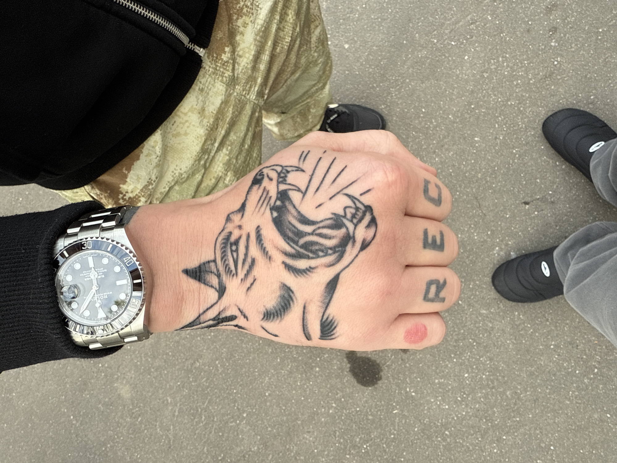 The image shows a person's hand with tattoos of a roaring animal on the back of the hand and the letters "RE" on the fingers, with a partially visible "C" on the fourth finger. A silver wristwatch is visible on the wrist. The person is wearing a black jacket and khaki pants. A pair of black shoes are visible nearby. The image was taken outside on a grey surface. There is no data to confirm or deny the location.