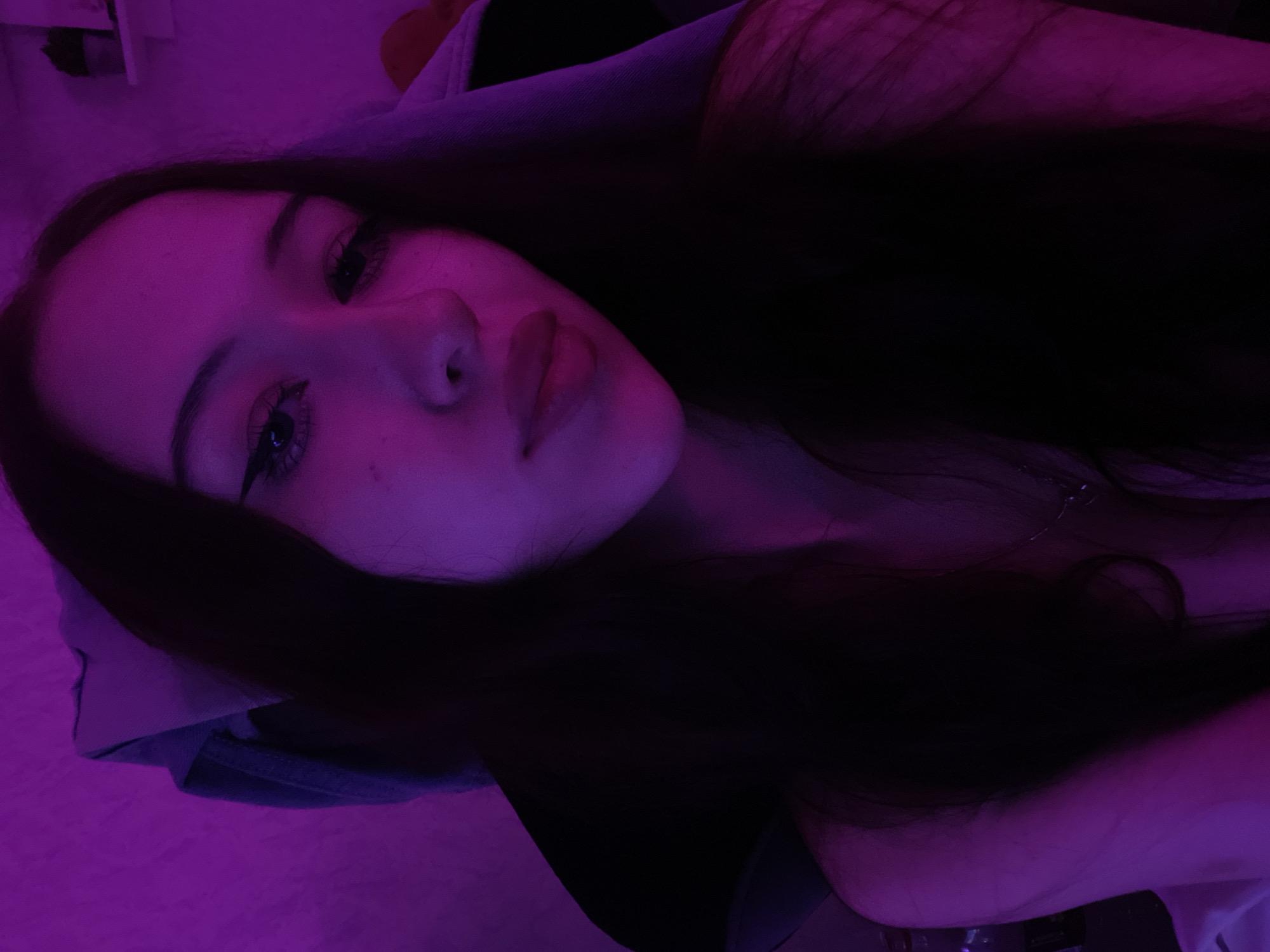 A young woman with dark hair and fair skin is shown in a close-up, self-taken photograph, lit by purple lighting. She is looking towards the camera with her eyes open and has winged eyeliner. Her lips are slightly parted. The background is dimly lit, with hints of clothing or fabric visible. There is no discernible indication of the location being in Votkinsk, Russia, or any specific urban setting. The image focuses solely on the woman's face and upper body.