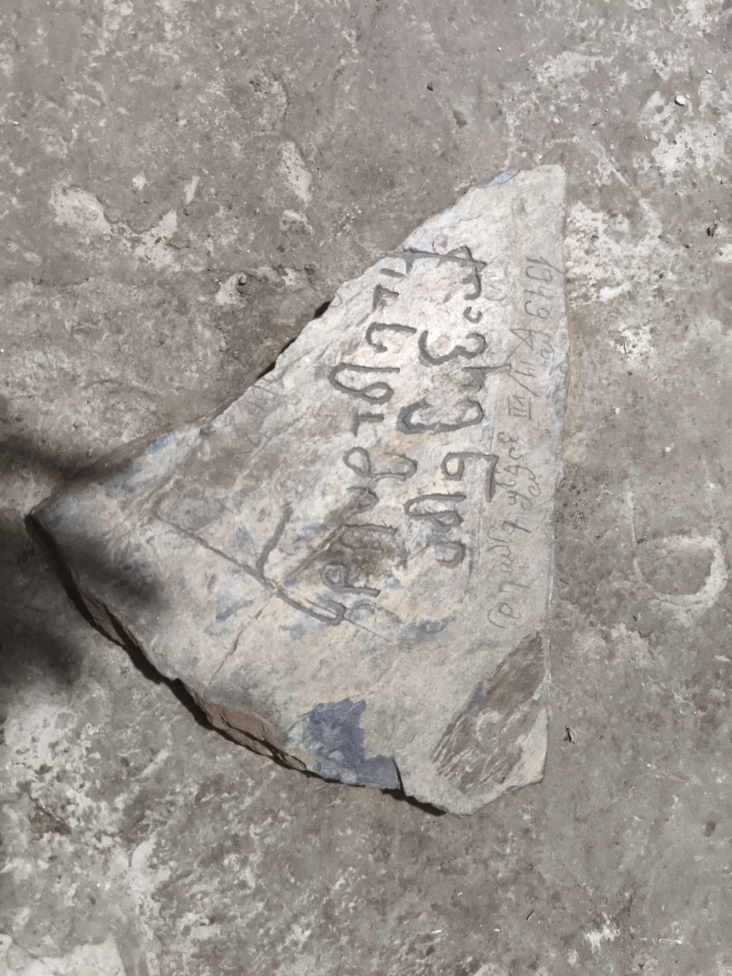 A triangular, weathered stone fragment, approximately 30 cm in length, rests on a rough, concrete surface. The stone exhibits faint markings, possibly inscriptions, in a dark gray pigment. Some of these markings appear to be numbers and what could be letters or symbols. On the right side of the stone, the numbers "1949" are clearly visible, followed by a character that resembles a reversed "R" or Cyrillic "Я", and then a few more unclear symbols. To the left of "1949", there are additional markings that are difficult to decipher. The concrete surface is gray and appears worn, with areas of lighter and darker discoloration and small pebbles embedded within it. The lighting suggests an indoor or shaded outdoor environment, casting a subtle shadow on the left side of the stone. The overall scene depicts an artifact found on a common, utilitarian surface.