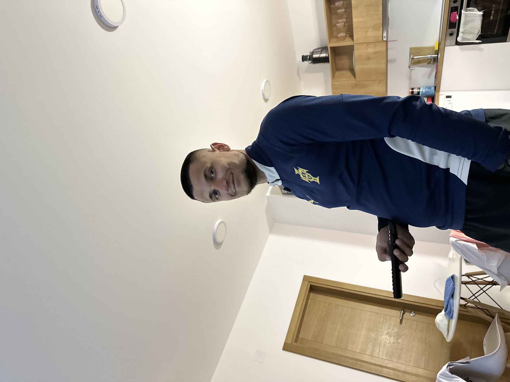 A young Caucasian male with short hair and a beard stands in a bright, modern kitchen in Donji Pobori, Montenegro. He is holding a black remote control in his right hand and is looking towards the camera with a slight smile. He is wearing a navy blue long-sleeved shirt with a light blue collar and a yellow crest on the left chest. Behind him, a wooden door is visible, along with a small round table with chairs and kitchen cabinets and appliances. The room appears to be a residential setting, possibly an apartment.