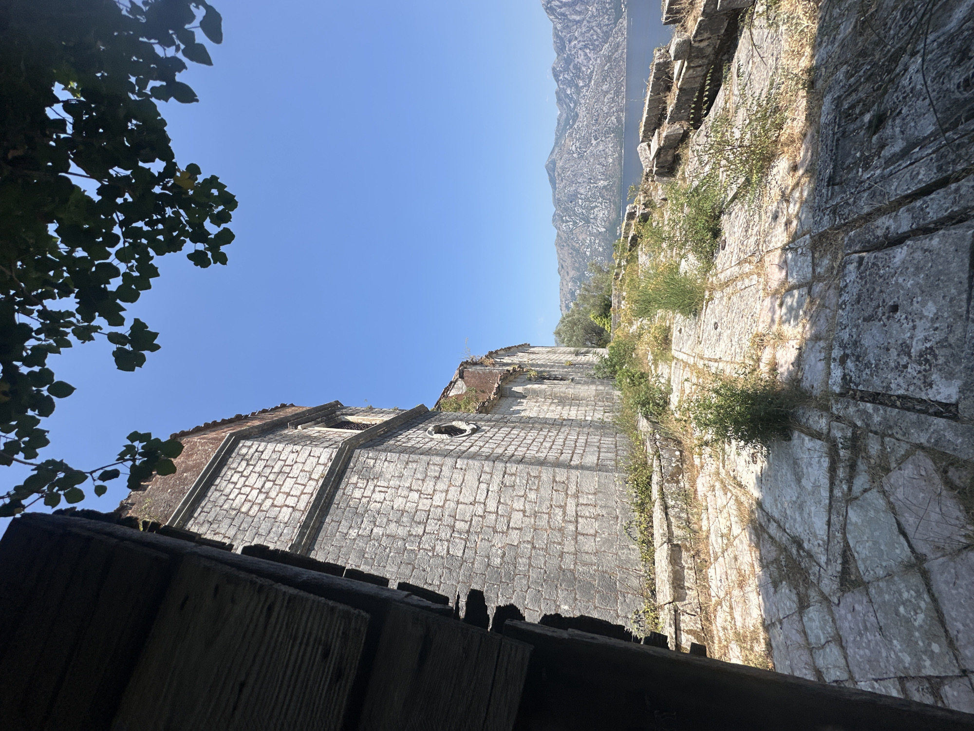 The image shows a view from the walls of a ruined stone building, possibly a church or fortress, overlooking the Bay of Kotor in Prčanj, Montenegro. In the foreground, a weathered stone pathway is partially obscured by shadows cast by an unseen structure. The pathway is made of large, irregular flagstones, with sparse vegetation growing between them. To the left, a portion of a dark, wooden beam or structure is visible.
The stone building itself is constructed from rough-hewn, grey stone blocks, showing signs of significant decay and weathering. A circular window is visible on its facade. The roof appears to be partially collapsed, with some red roofing tiles visible. Greenery and vines are creeping up the sides of the structure.
Beyond the building, the calm, blue water of the bay stretches out, meeting a backdrop of rugged, mountainous terrain. The mountains are steep and rocky, with patches of scrub and trees on their slopes. The sky is a clear, bright blue. The scene suggests a historical site with a commanding view of the natural landscape.