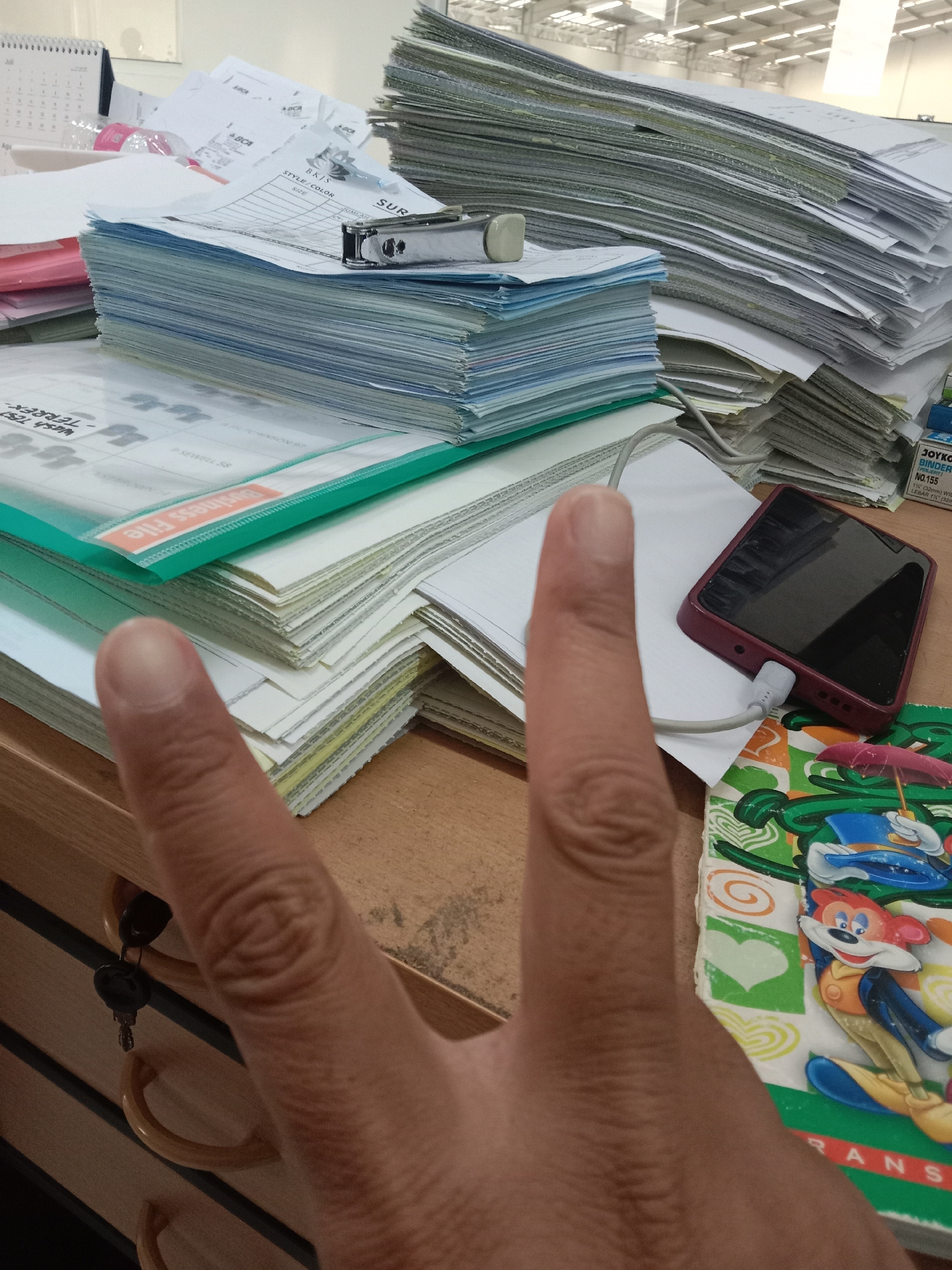 A close-up, eye-level, medium shot captures a desk cluttered with documents and office supplies. In the foreground, a person's left hand is positioned with the index and middle fingers extended in a peace sign, obscuring a portion of the desk and objects behind it. The desk surface, made of wood, is visible in the lower half of the frame and shows signs of wear. On the desk, a stack of blue and white papers is secured by a silver stapler. To the right of the papers, a dark red smartphone is connected to a white cable and rests on a colorful paper or folder featuring cartoon characters. Behind the main stack of papers, a much larger, disorganized pile of documents is present, extending towards the upper right corner of the image. A desk calendar with the month "July" visible and a clear plastic water bottle are positioned on the left side of the desk, behind the initial stack of papers. Keys hang from a drawer pull on the lower left side of the frame. The background is out of focus, but appears to be an indoor environment with office lighting and ceiling structures, potentially a warehouse or large office space, but this is not definitively identifiable as Semarang, Indonesia.