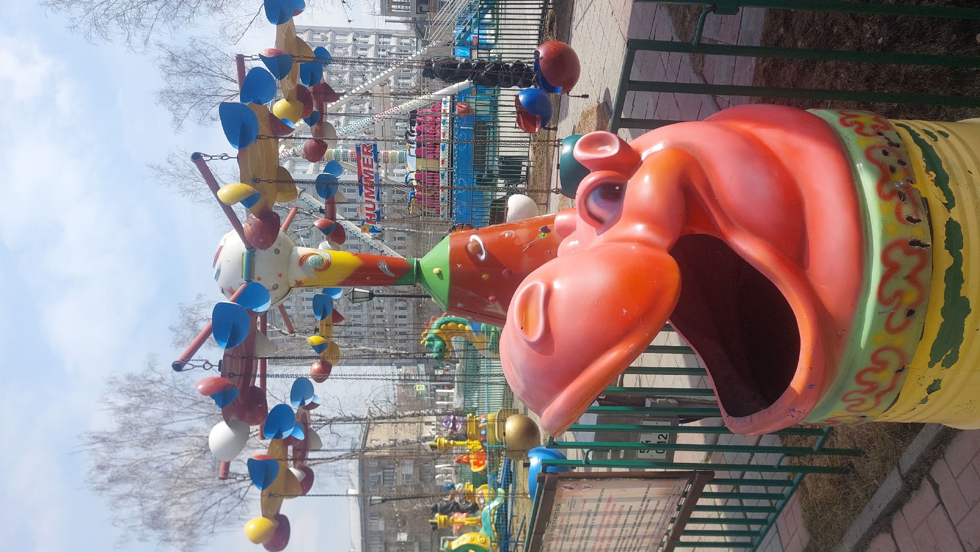This is an outdoor shot in Novosibirsk, Russia, focusing on amusement park rides. The prominent feature is a large, red and orange hippopotamus-shaped seat with a gaping mouth. Adjacent is a colorful, spinning ride adorned with whimsical shapes like leaves and balls. In the background, there is a Ferris wheel and buildings of the city. The ground is paved with bricks, and the scene is framed by greenery.