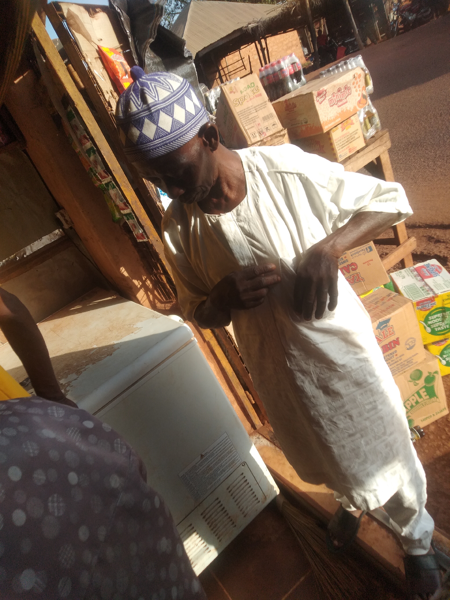 In Ejule, Nigeria, an elderly man wearing a patterned blue and white cap and a long, white garment stands next to a refrigerator in front of a small outdoor shop. The shop displays cardboard boxes containing various products, including drinks and snacks. The ground is reddish-brown. The lighting suggests it is daytime. A partial figure of a person wearing a patterned garment is also visible next to the refrigerator. A broom is on the ground next to the man.