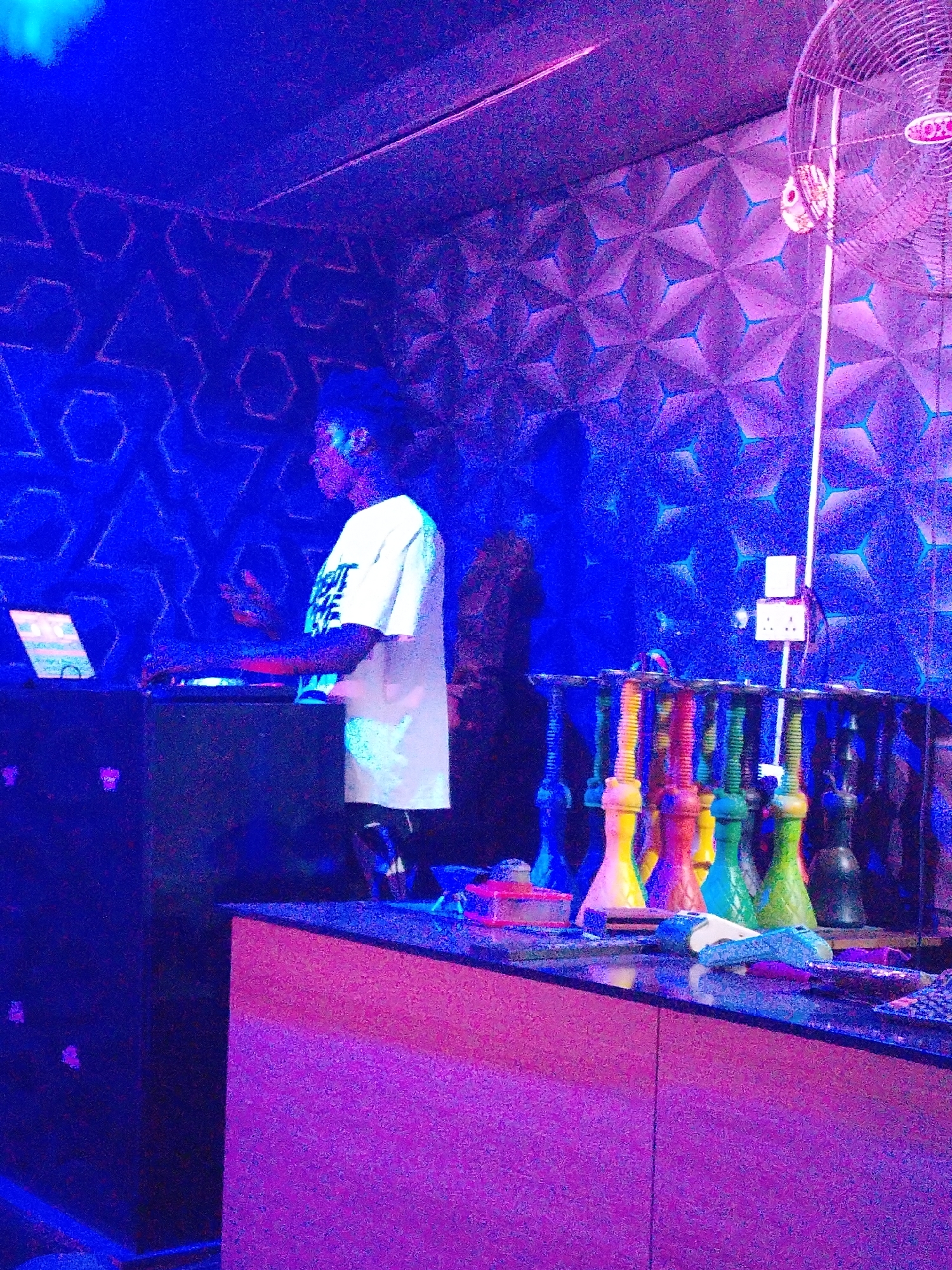 The image depicts a scene within a club, likely a hookah bar, in an unknown city. A person is standing behind a counter working on a DJ setup with a laptop and mixer. Several hookahs are lined up on the counter. In the background, a wall with a geometric pattern and a fan are visible. The lighting is predominantly blue and purple, creating a club atmosphere.