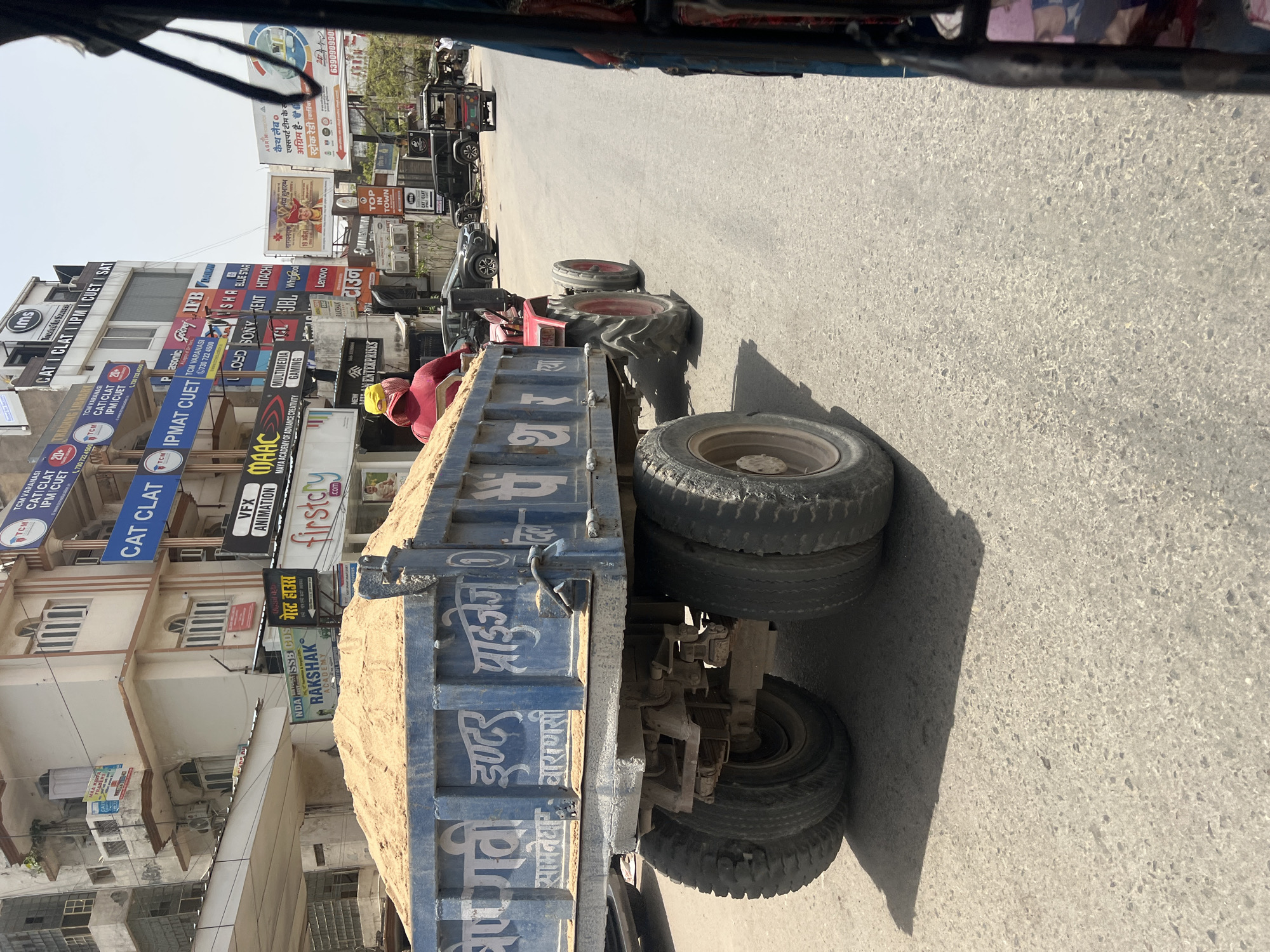 A medium-angle shot looking out from a vehicle such as an auto-rickshaw shows a blue truck carrying a load of sand on a road in Varanasi, India. The truck is to the right, with its rear tires and the truck bed heavily featured. Next to the truck is a red tractor. Across the street is a building covered in a dense array of commercial signage for various businesses, including test preparation services ("CAT CLAT IPM CUET SAT"), electronics (Panasonic, Sony, Kent, Hitachi, etc.) animation/VFX, and others. Further down the road are parked vehicles, including auto-rickshaws and cars. The sky is clear and a light blue color.