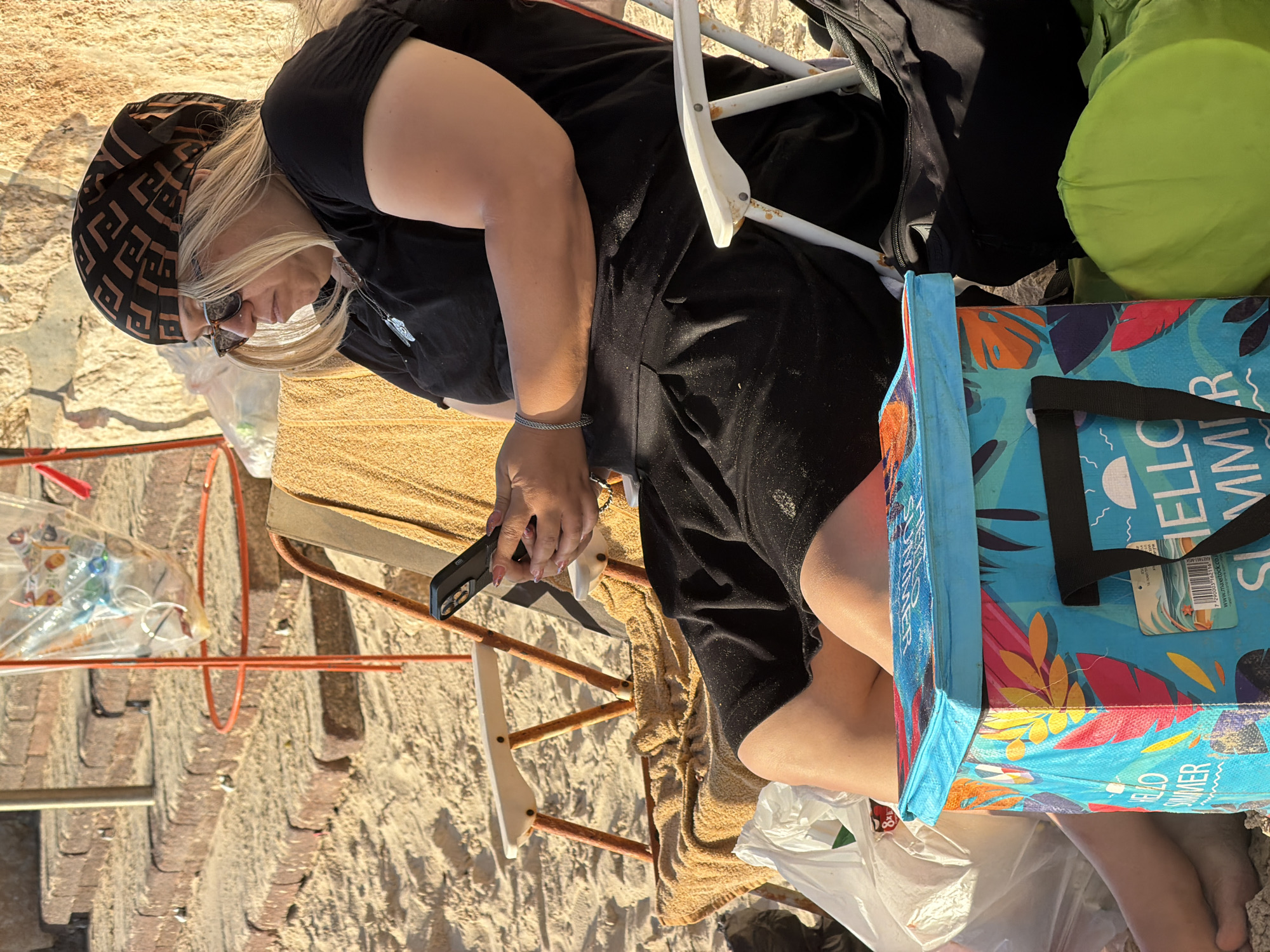 The image shows a woman sitting on a beach chair on a sandy beach. She is wearing a black t-shirt, a black skirt, sunglasses, and a patterned headscarf. She is holding a smartphone in her hands and appears to be looking at it. Next to her is a colorful cooler bag with the words "HELLO SUMMER" printed on it. There are also some plastic bags and a green inflatable object visible. In the background, there are stone steps and a wall, suggesting an urban beach setting. The lighting indicates it is daytime, likely afternoon or late afternoon, given the warm tones of the sunlight. The sand is lightly textured, with some small debris present. The overall scene depicts a casual day at the beach, possibly in Tel Aviv, Israel, known for its urban beaches.