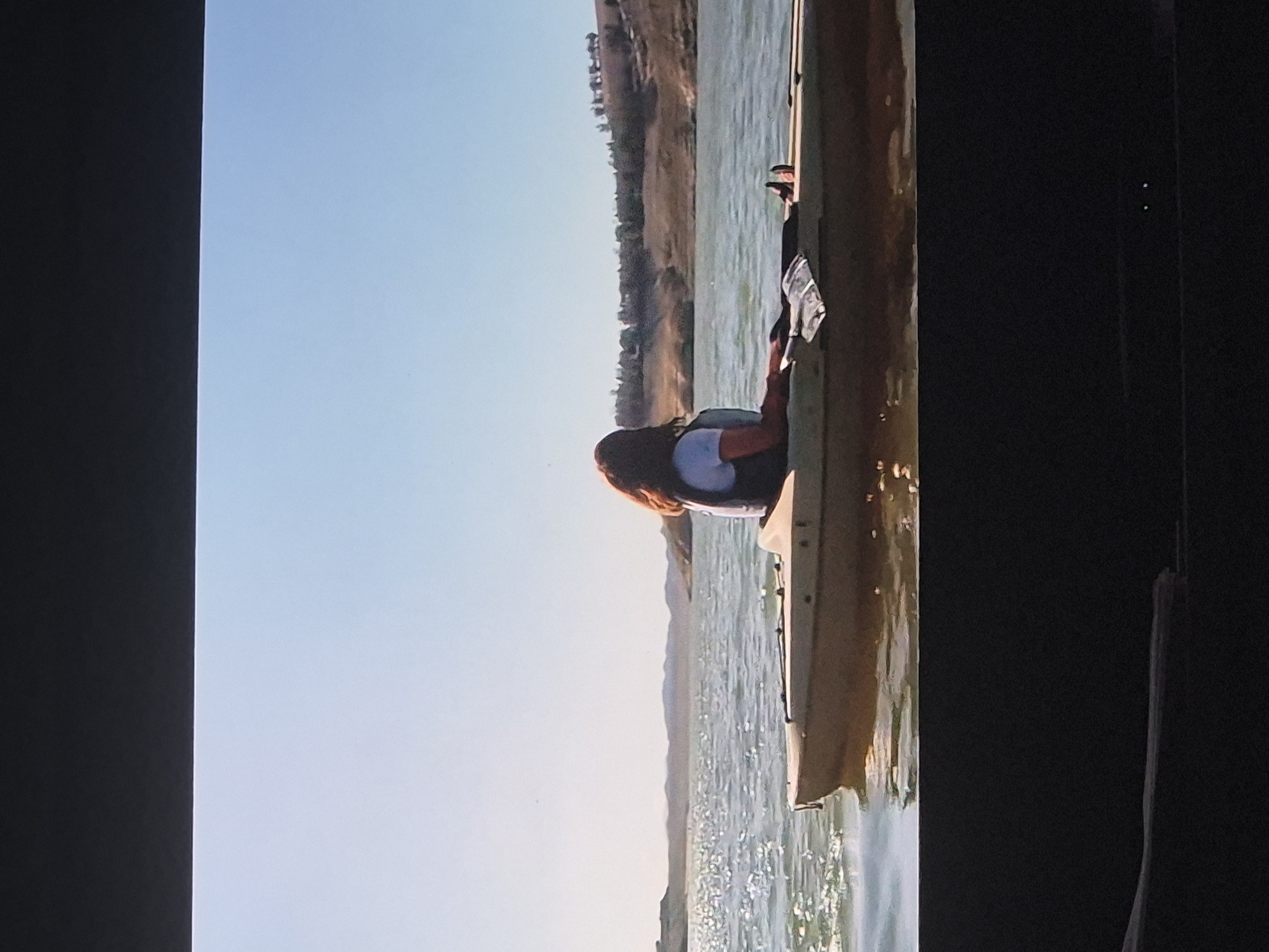 The image displays a woman in a boat on a body of water under a clear sky, appearing to be a photograph of a screen.
**Subjects:** A woman with long, reddish-brown hair is visible from the chest up, positioned in a light-colored boat, likely a kayak or canoe. She is wearing a light-colored top and a dark life vest or backpack, with her back towards the camera and her head turned to the left. The boat is beige or tan. In the mid-ground, a expansive body of water is seen, reflecting sunlight. A distant shoreline with undulating hills covered in sparse, dry vegetation forms the background. The upper portion of the image is dominated by a bright, clear sky.
**Activity or event taking place:** The woman is seated or reclining in the boat, suggesting a recreational activity such as kayaking or boating on the water. Her posture indicates a moment of relaxation or observation of the surroundings.
**Notable details:** The time of day appears to be late afternoon or early morning, characterized by bright, soft sunlight casting reflections on the water. The weather is clear and sunny. The distant shoreline suggests an arid or semi-arid geographical location with natural, undeveloped land. The visible reflections on the water indicate a calm surface. The entire image seems to be a photo taken of a screen, given the black bars on the sides and slight screen artifacts.
**Visible text:** There is no visible text in the image.