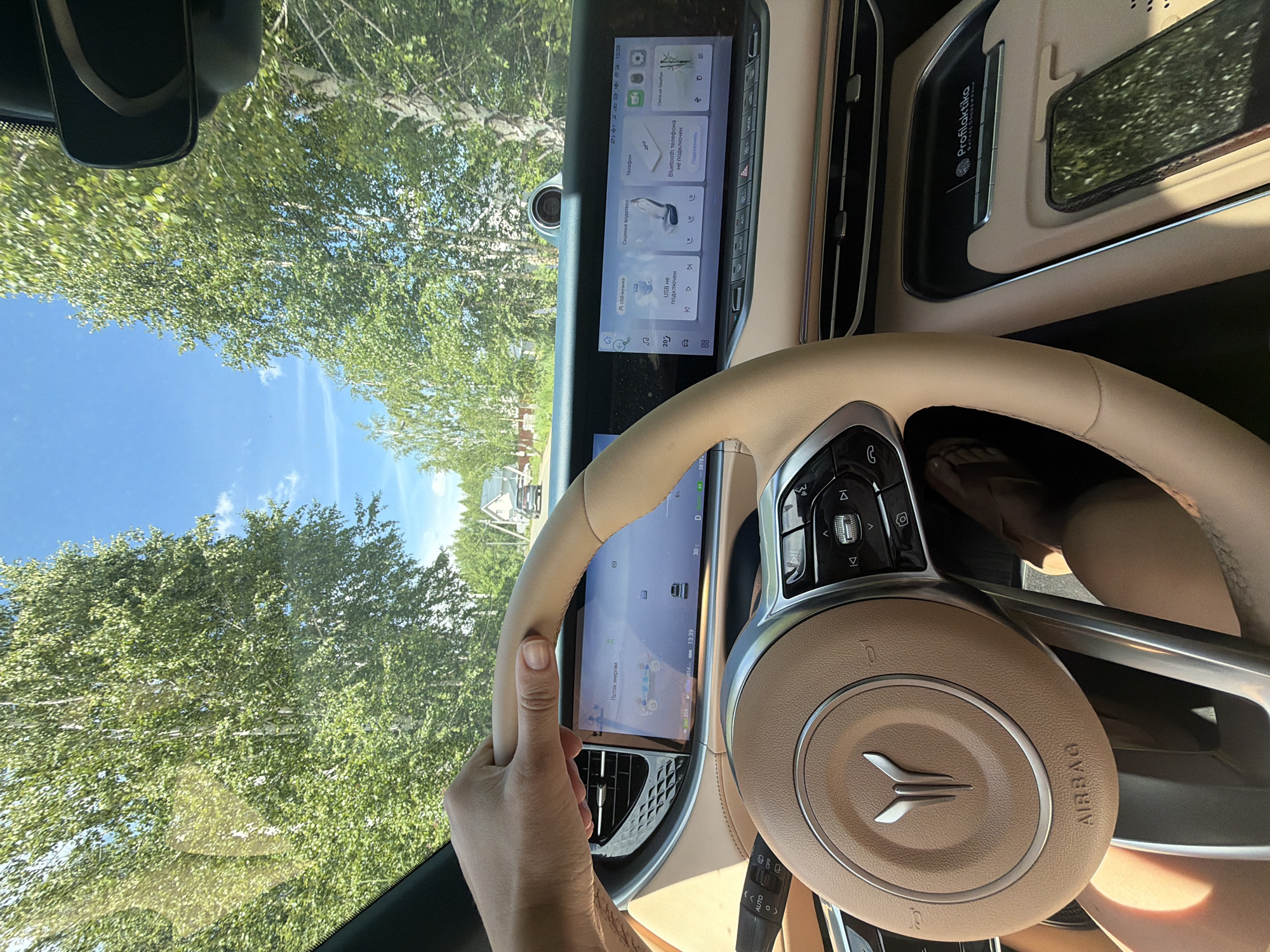 A first-person perspective shot captures a hand on the steering wheel of a car. The interior of the car is visible, featuring a large digital display dashboard and a tan leather steering wheel with an emblem. The car is on a road lined with trees, and houses are visible in the distance. The sky is blue with scattered clouds. The car appears to be traveling through a rural area, possibly on a summer day, given the lush greenery. The scene suggests a typical driving experience in a suburban or rural setting in Malinovka, Russia.