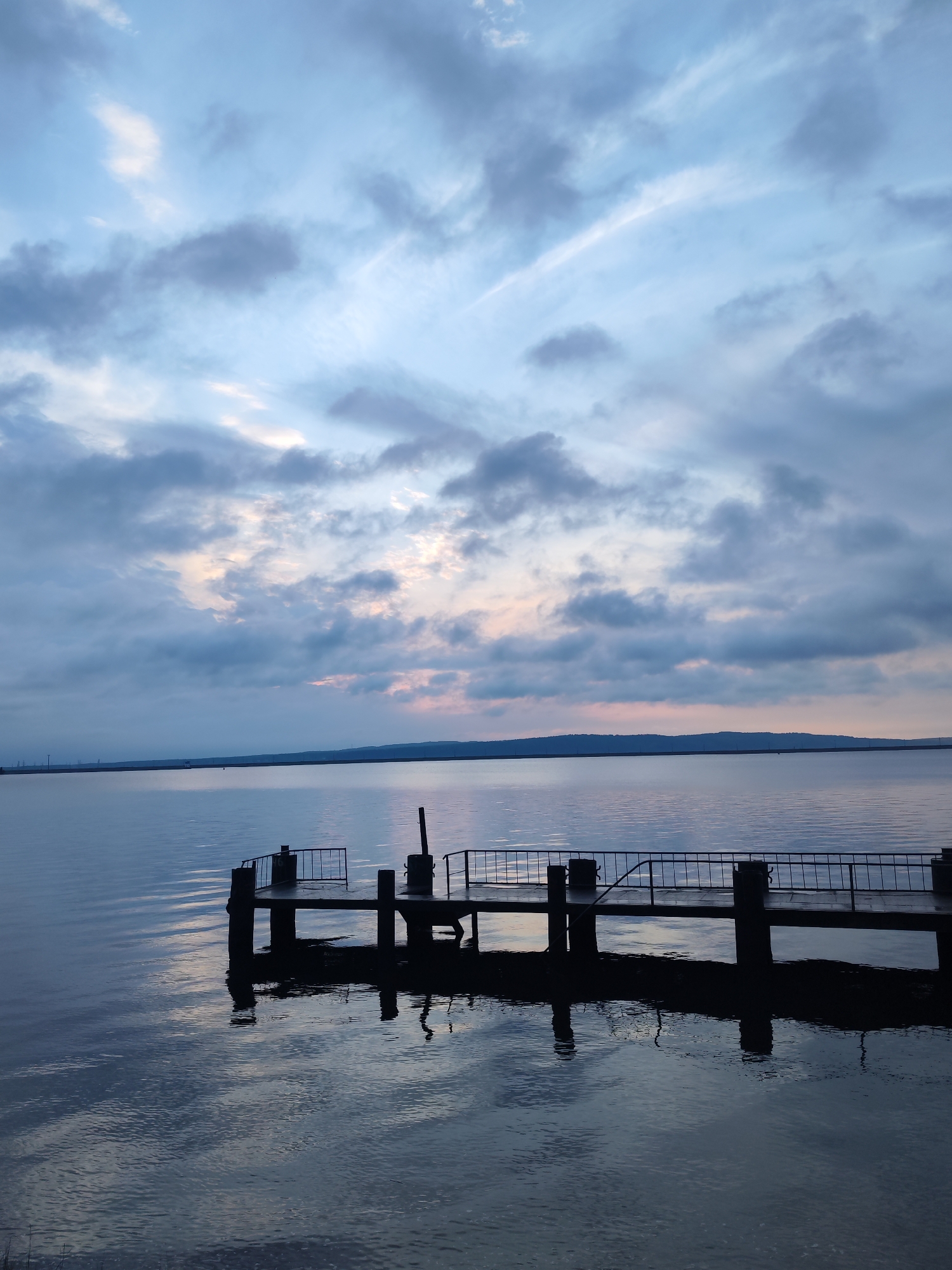 The image shows a tranquil scene at a lake or large body of water under a cloudy sky. The time of day appears to be either dusk or dawn, given the soft light and coloration in the sky. A small, dark pier or platform juts out into the water, supported by pilings. A railing is visible along one side of the platform. The water is calm with slight ripples and reflects the sky and the pier. The horizon includes a darker strip of land, possibly a distant shore or island. The image was taken at Lake Saygatka in the city of Chaikovsky, Russia.
