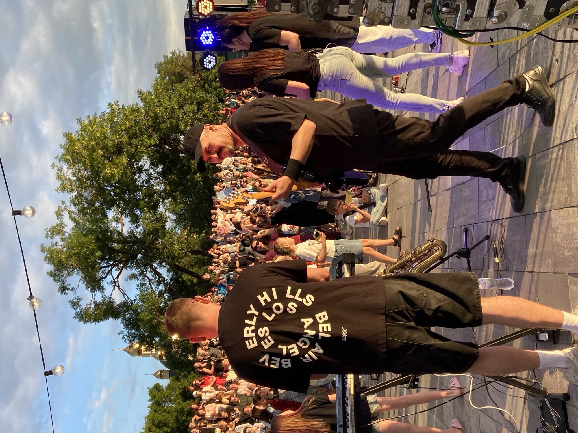 The image captures a live outdoor performance, likely at a festival or concert, in the city of Unknown, Unknown. In the foreground, a man wearing a black t-shirt with white circular text, black shorts, and white socks is seen from behind, facing away from the camera. He is standing in front of a keyboard. To his right, a man with a black baseball cap and black t-shirt is playing a guitar and looking towards the camera. He has a mustache. Behind these two men, a crowd of people are gathered, watching the performance. In the background, there are trees, a building with golden domes resembling a church, and stage lighting equipment. Several string lights are suspended above the scene. The overall atmosphere is that of an evening outdoor event, with the setting sun casting a warm glow.