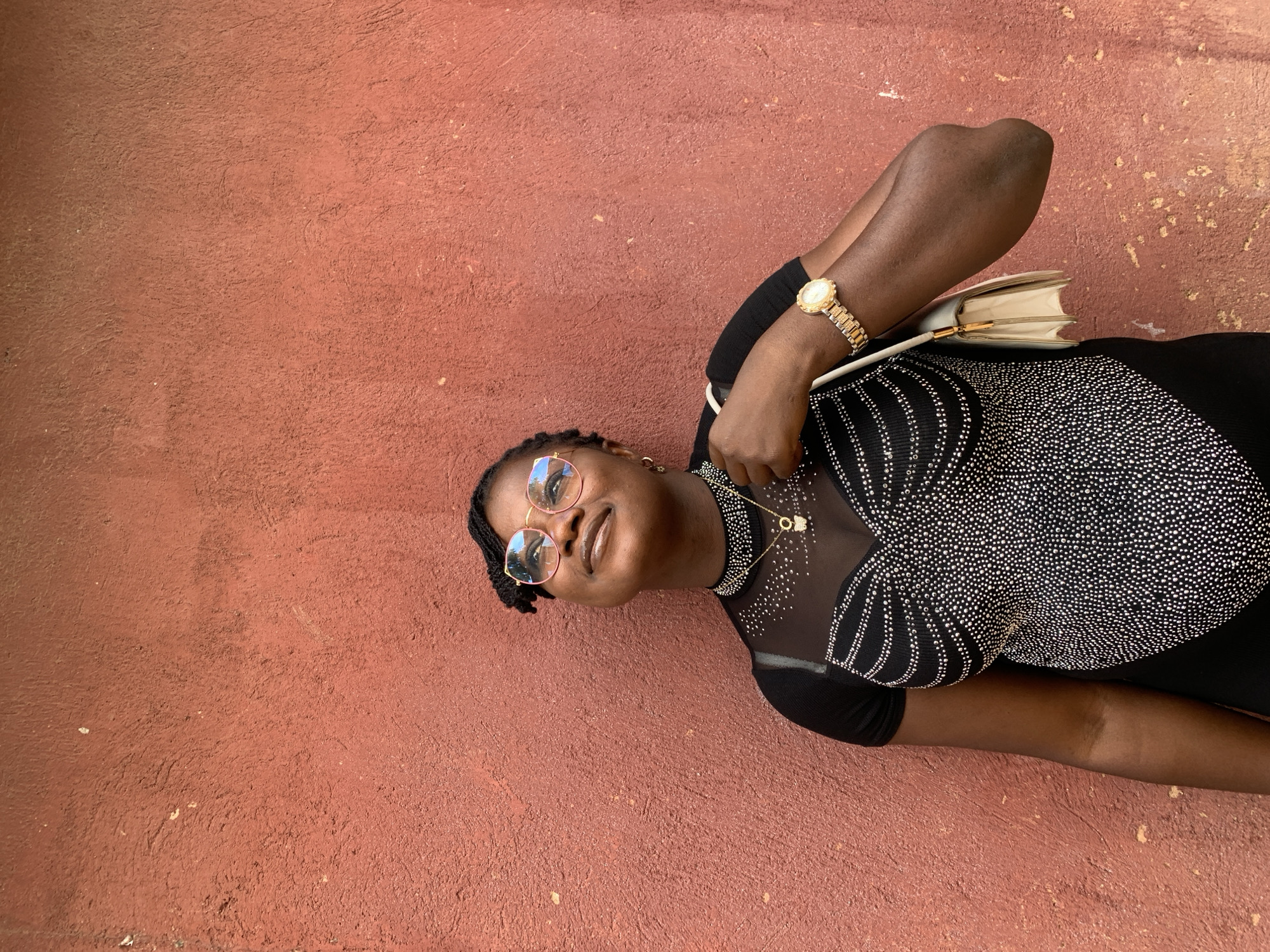 A young woman poses against a textured, terracotta-colored wall. She is wearing a black, sheer-necked top adorned with silver embellishments, gold watch, and round, rose-tinted glasses. She also wears a delicate necklace. She carries a light-colored, structured handbag. The background is a plain wall, suggesting an urban setting. The location in the city of Jos, Nigeria is not directly discernible from the image.