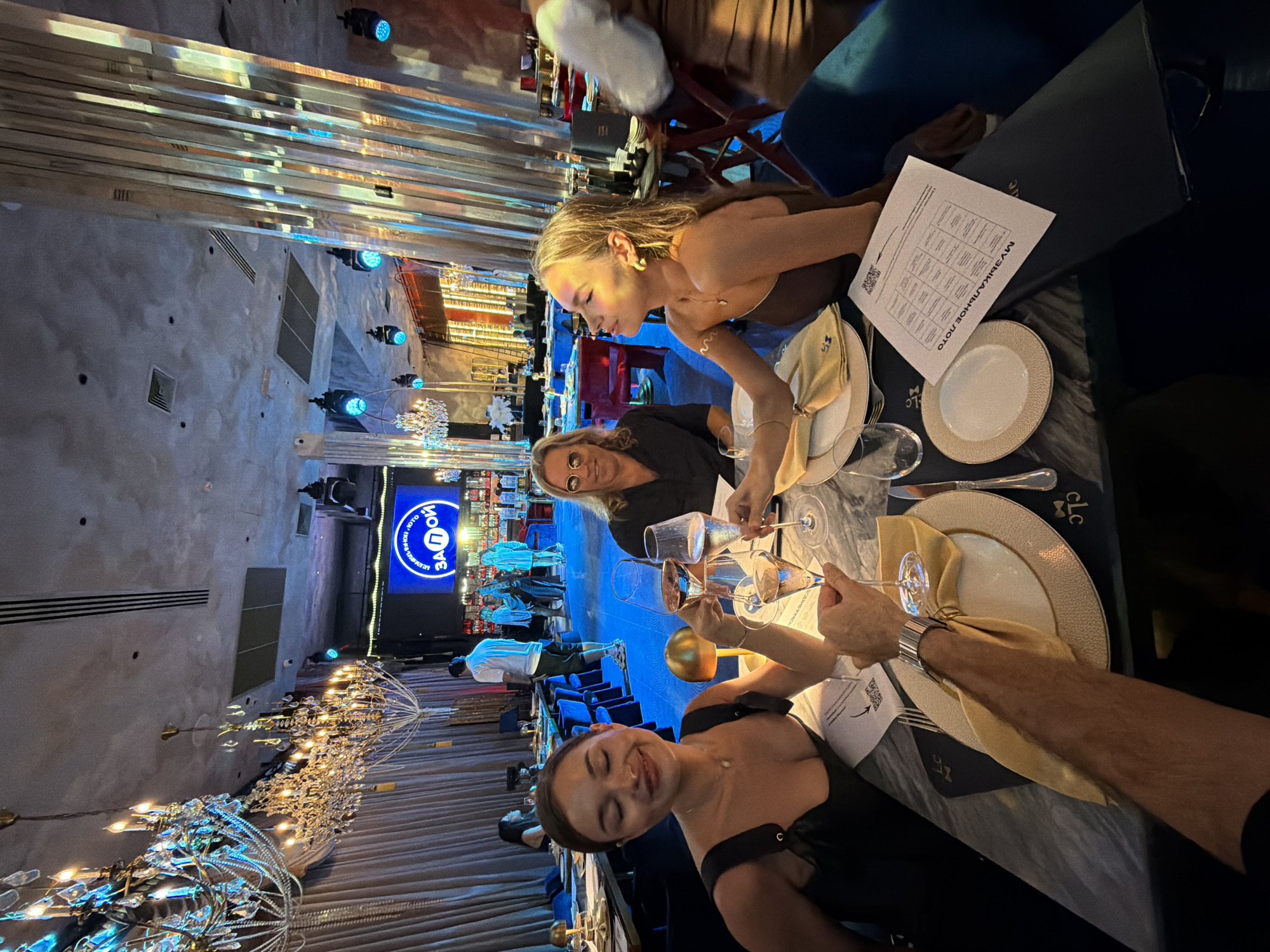 The image shows a group of three women and one arm of a man toasting with champagne flutes at a dinner table inside a restaurant in Choeng Thale, Thailand. The women are sitting at the table, while the man's arm extends into the frame for the toast.
The restaurant is decorated with elaborate chandeliers and illuminated metal structures on the ceiling. A large screen in the background displays what appears to be an event or advertisement in Russian. The tables are set with plates, napkins, cutlery, and menus. The table surface is dark and has a marble-like pattern. The restaurant is well-lit with both ambient and decorative lighting. The women are dressed in evening attire, suggesting a special occasion.