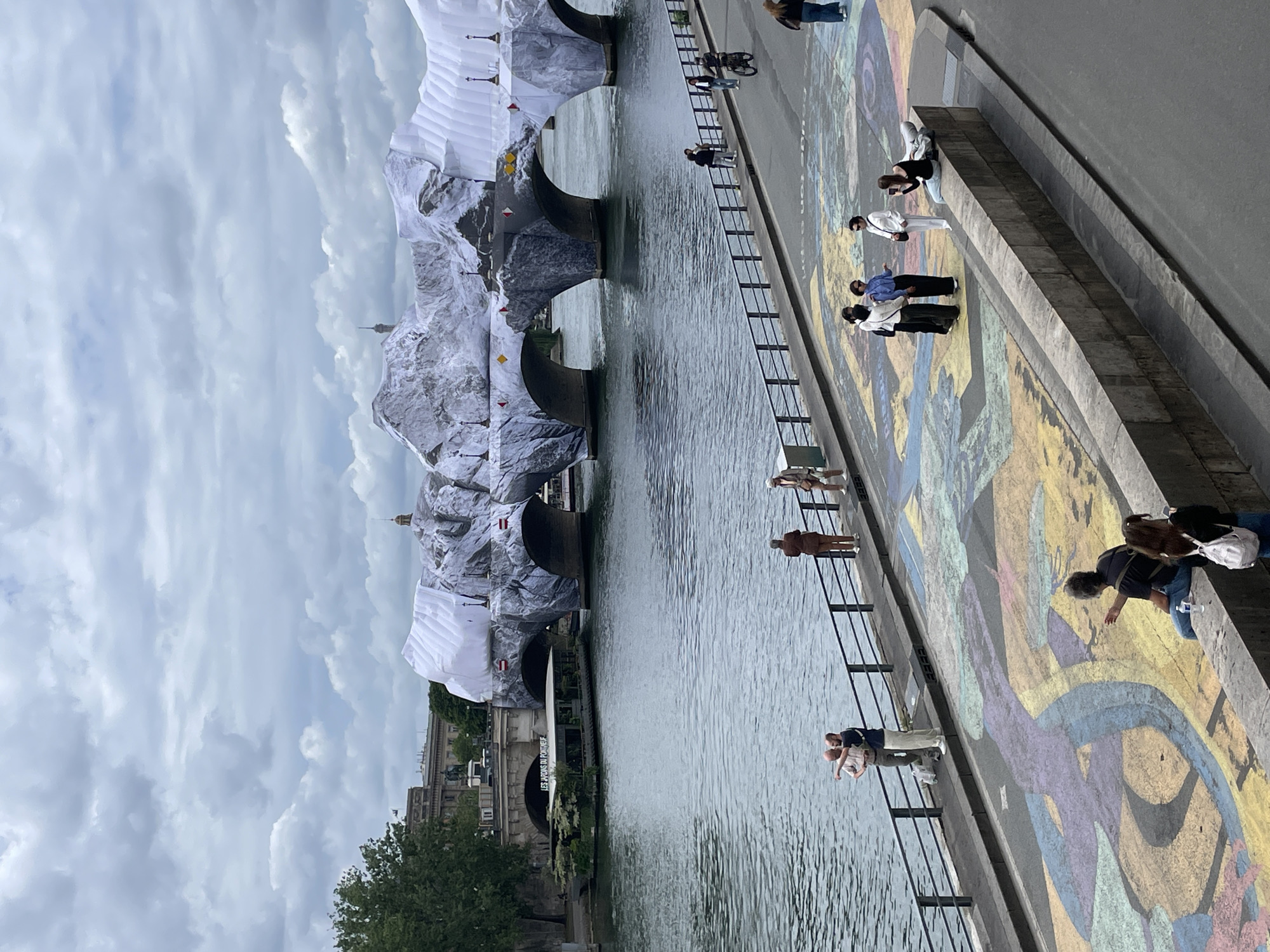 The image shows a view of the Pont Alexandre III bridge in Paris, France, draped with a large art installation resembling a snowy mountain range. The bridge spans a wide body of water, likely the Seine River, with a boat docked on the left bank. A promenade runs along the right bank of the river, parallel to the bridge, where several people are visible.
On the promenade, a group of individuals is walking, standing, and taking photographs. One man is riding a bicycle. The ground of the promenade features a colorful chalk art mural. Further to the right, more people are seated and standing. In the background, several buildings and structures are visible, including a dome and a tall, slender tower that resembles the Eiffel Tower. The sky is overcast with clouds.