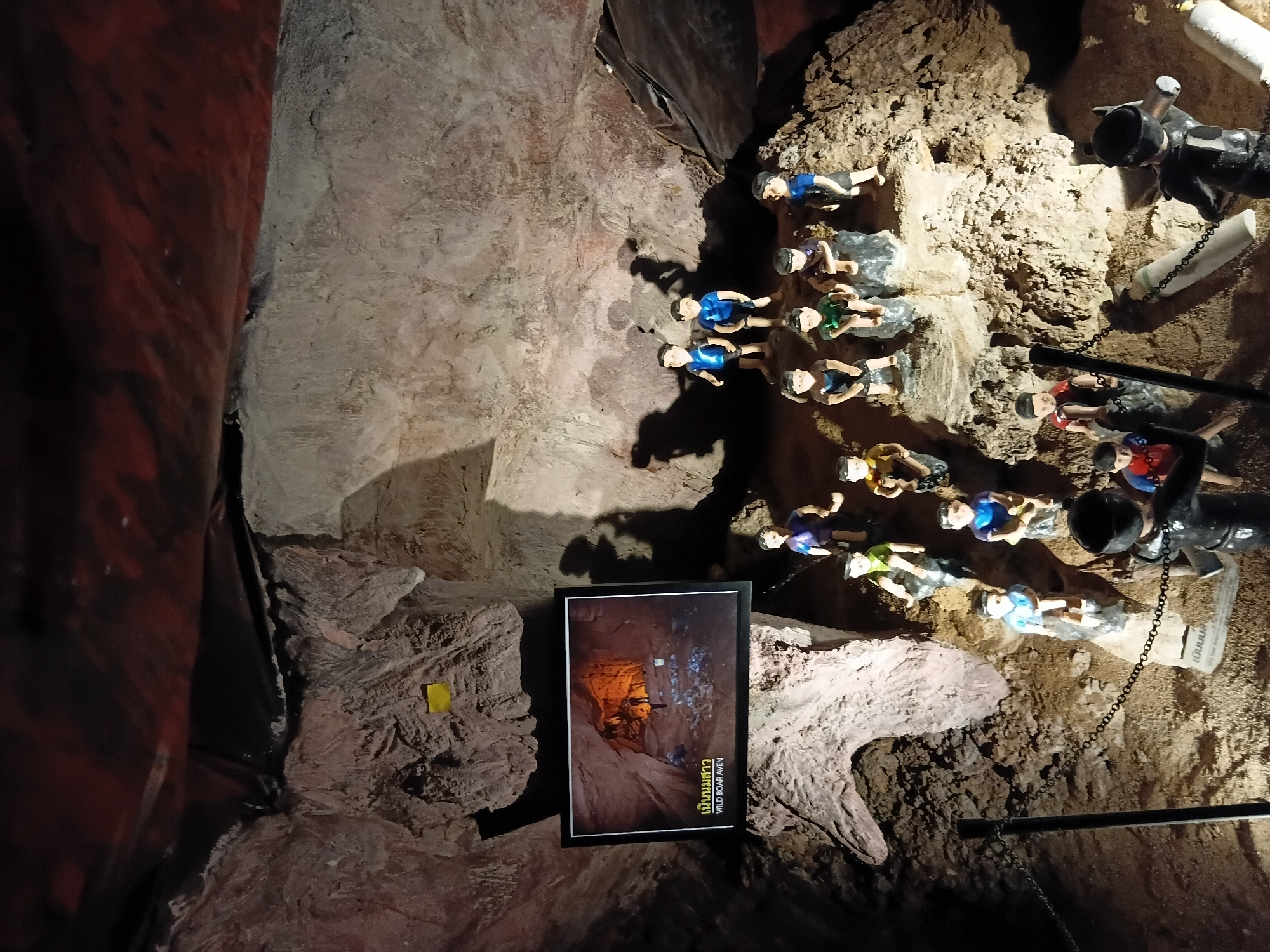 The image depicts an interior scene of a cave, likely a tourist attraction, in Mae Sai District, Thailand. The cave walls are rough and textured, illuminated by artificial lighting that casts shadows. In the foreground, a series of small, human-like figurines are arranged on ledges and the cave floor. These figurines appear to represent children and are dressed in clothing. Some figurines are seated, while others are standing. They are positioned in small groups.
To the left of the figurines, a framed digital display hangs on the cave wall. The display shows an image of the interior of a cave, with text in Thai script above the English words "WILD BOAR AVEN".
A black chain fence runs across the lower portion of the image, separating the viewer's perspective from the figurines and the cave display. The ceiling of the cave is visible at the top, with a dark, possibly tarpaulin or plastic covering creating a ceiling effect. The overall impression is a staged exhibit within a natural cave environment, designed to tell a story or commemorate an event, likely related to the "Wild Boar" incident.