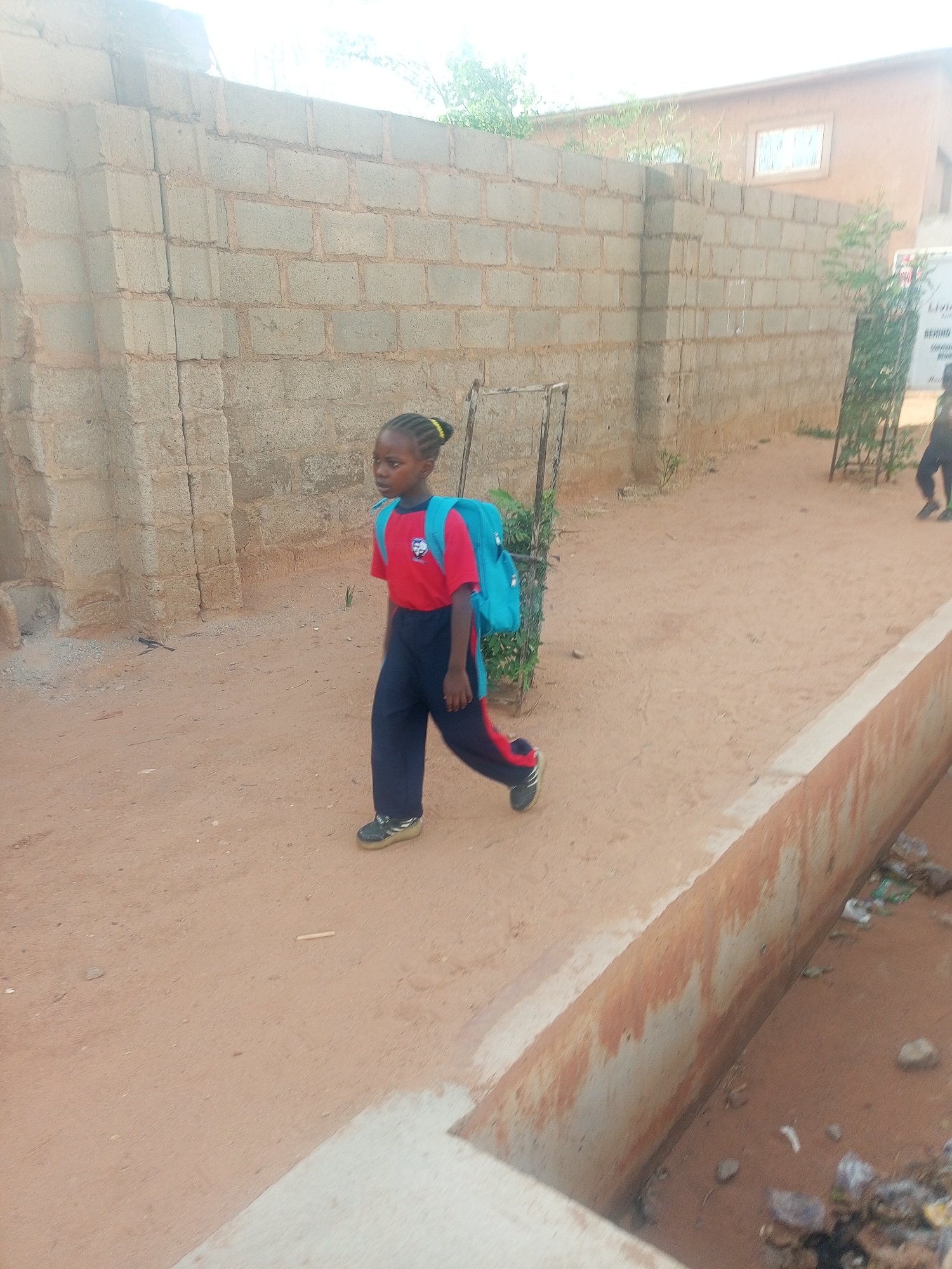 A young Black girl wearing a red short-sleeved shirt, dark blue pants, and black shoes with a light blue backpack walks on a dirt road. The girl is walking to the right in the image, and her gaze is directed forward. Behind her is a wall constructed of light gray cinder blocks. Further down the road is a taller building, also of cinder block construction. To the right of the dirt road is a low concrete wall, with what appears to be a drainage ditch beside it. The ditch appears to contain litter. The location is Unknown, Unknown.
