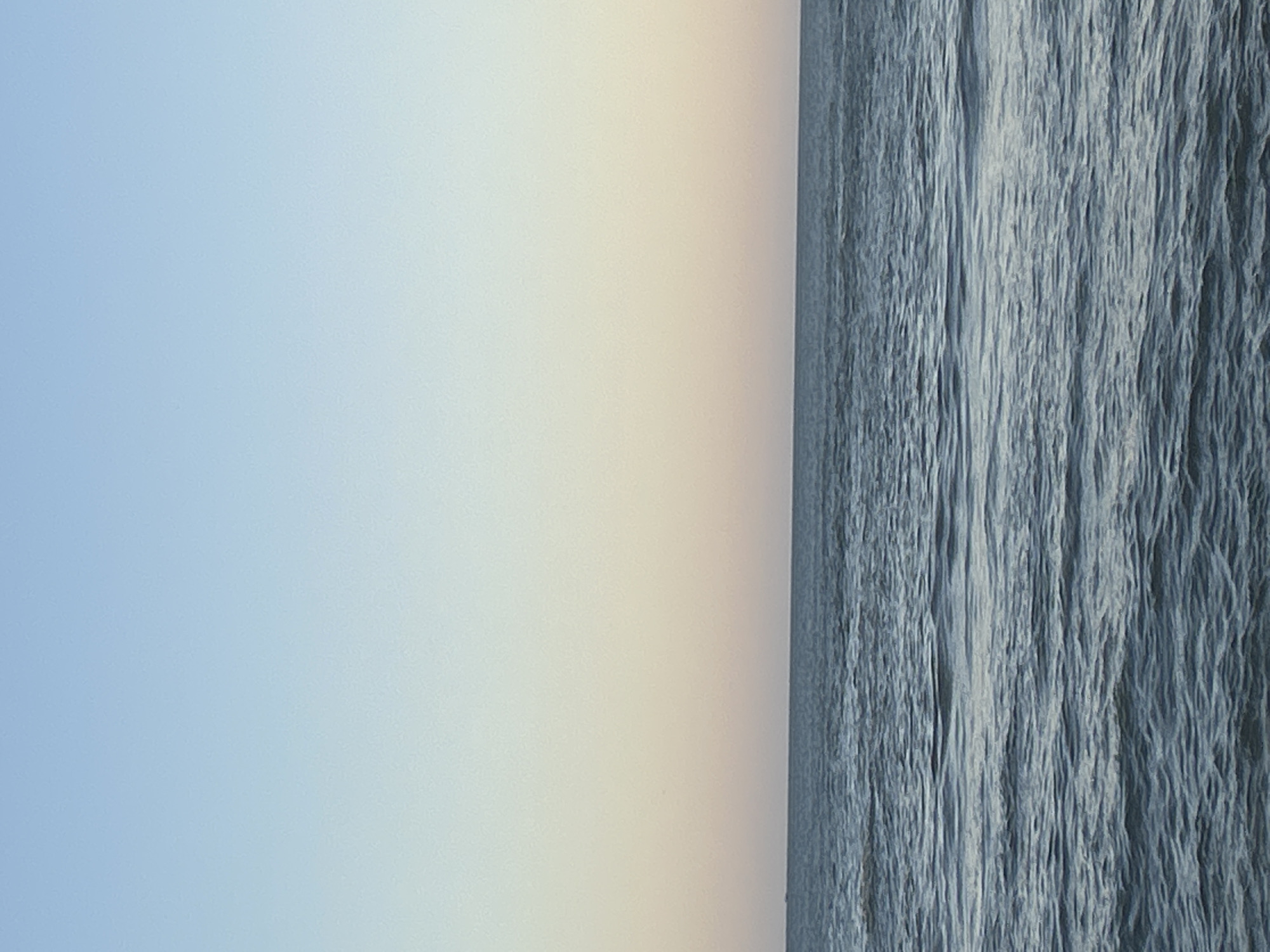 The image shows a division between a sky and a body of water, likely an ocean. The left portion depicts a soft blue sky transitioning to a pale yellow near the horizon. The right portion shows a textured surface of the water, displaying various shades of blue and grey. The water surface appears rippled or wavy. The image is oriented vertically. Without additional information, the location in Mazatlán, Mexico cannot be specified.