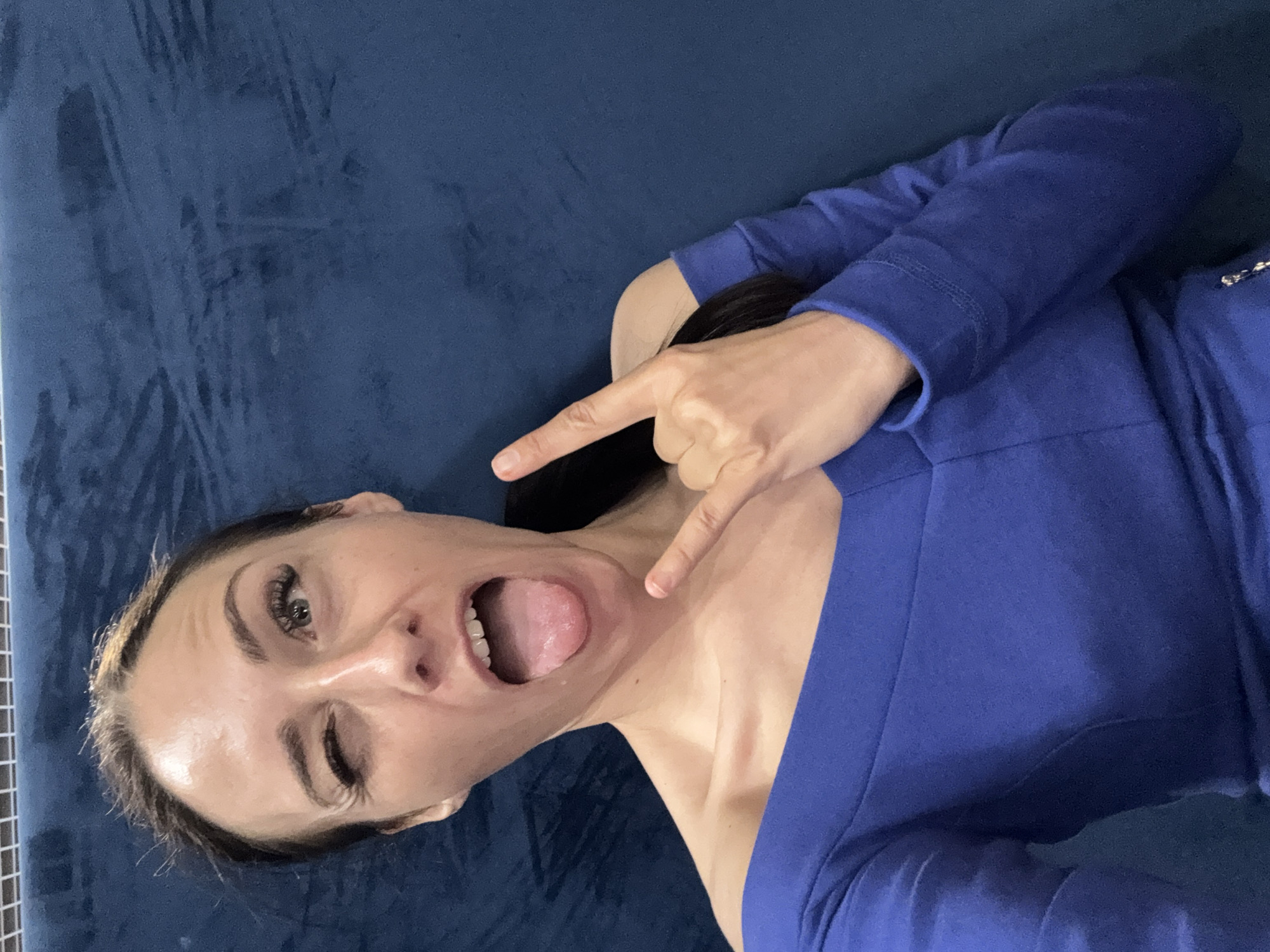 A woman is photographed from the chest up, against a blue, textured background. She is wearing a royal blue off-the-shoulder top. Her left eye is winked shut, and her tongue is sticking out. Her right hand is raised, with her index and pinky fingers extended in a rock and roll gesture. Her hair is dark brown and pulled back into a ponytail. The lighting is bright and appears to be from overhead. The background texture suggests it could be a sofa or a padded wall. The overall impression is playful and energetic. There is no discernible location in Saint Petersburg, Russia.